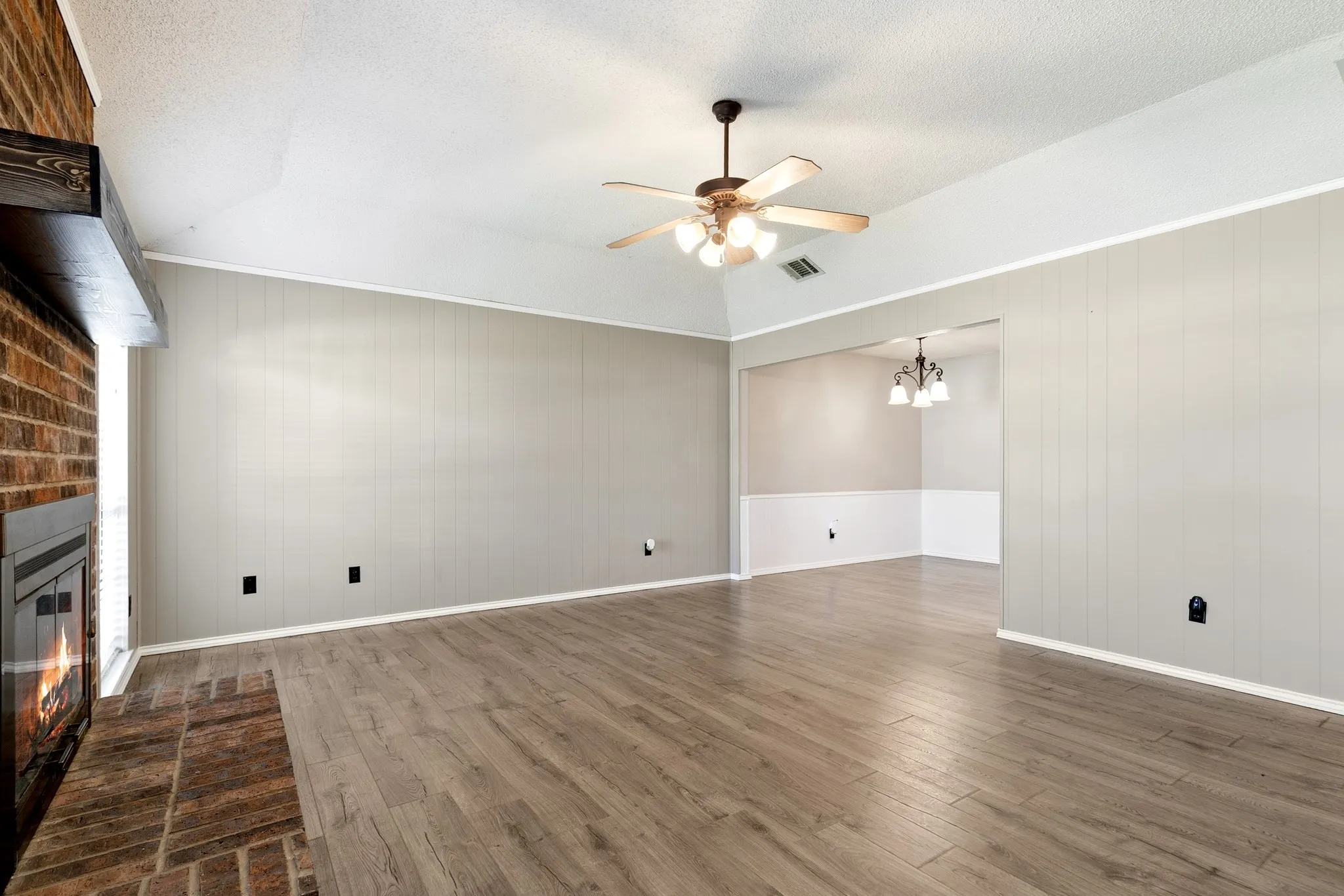 Spacious living area featuring wood-finish flooring, a brick fireplace with a wood mantle, and a ceiling fan with integrated lighting