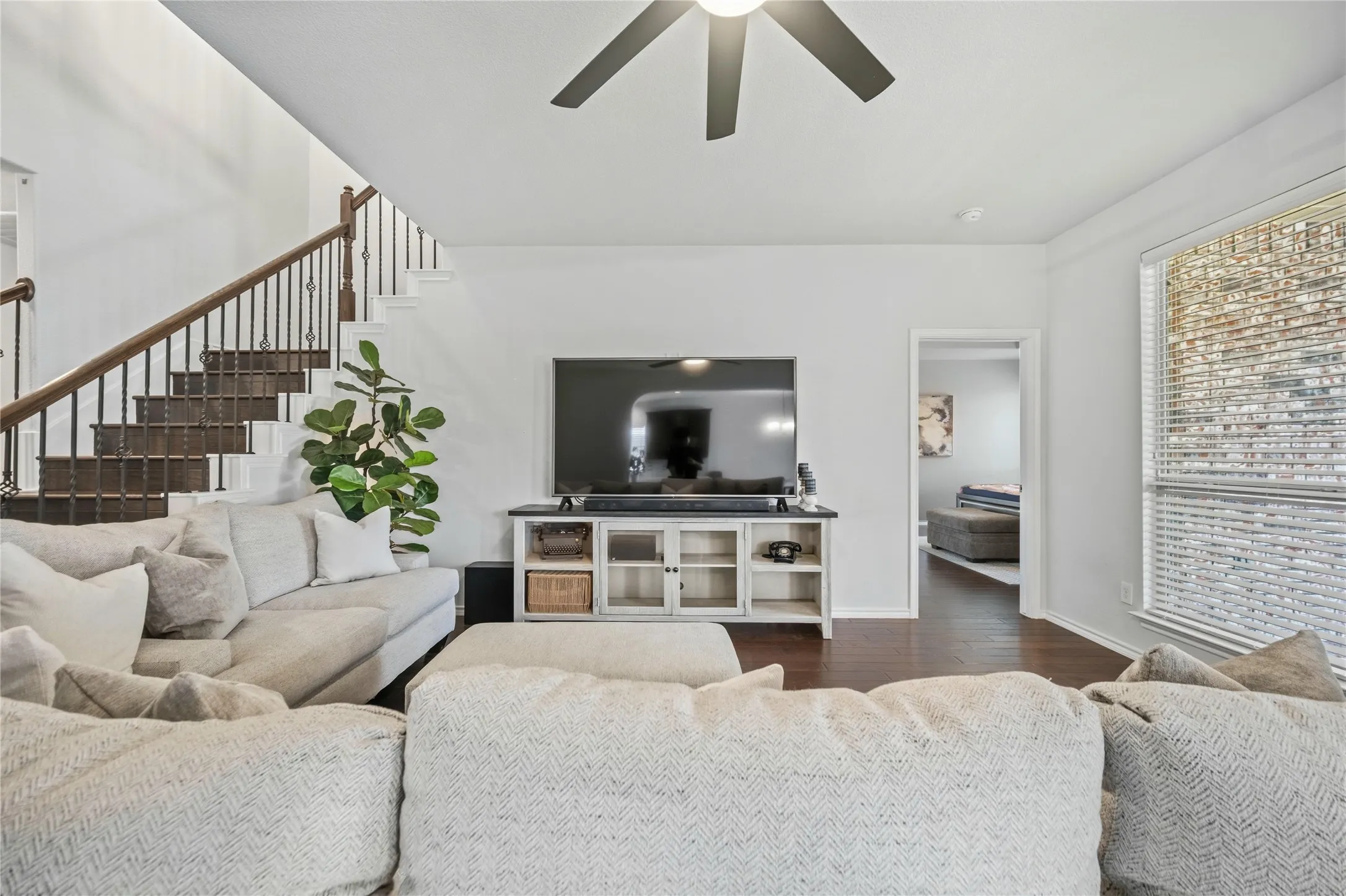 Spacious living area featuring wood-finish flooring, a contemporary ceiling fan, and a prominent staircase with dark wood treads and wrought iron balusters