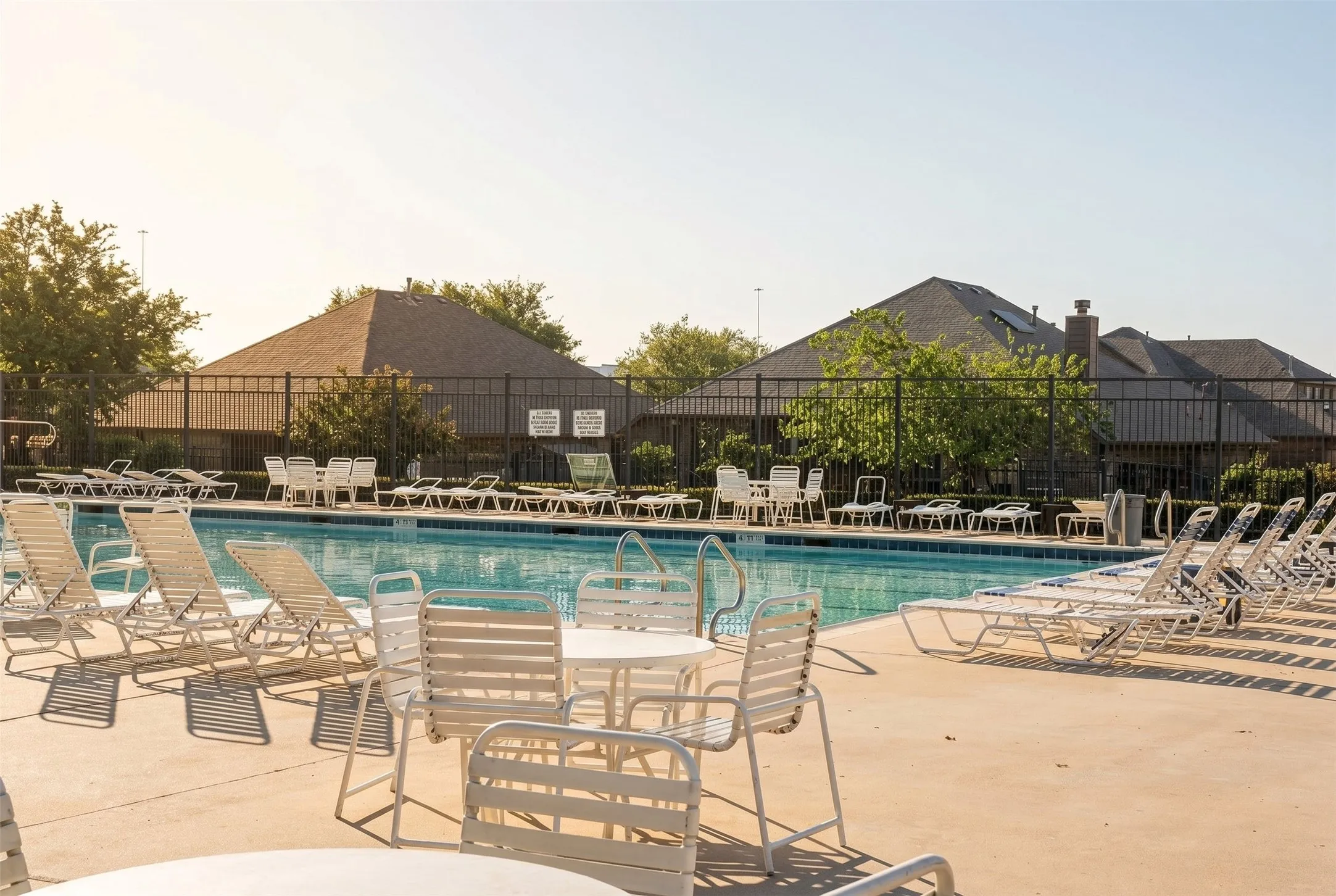 Community pool with a concrete surround, featuring multiple lounge chairs and poolside tables