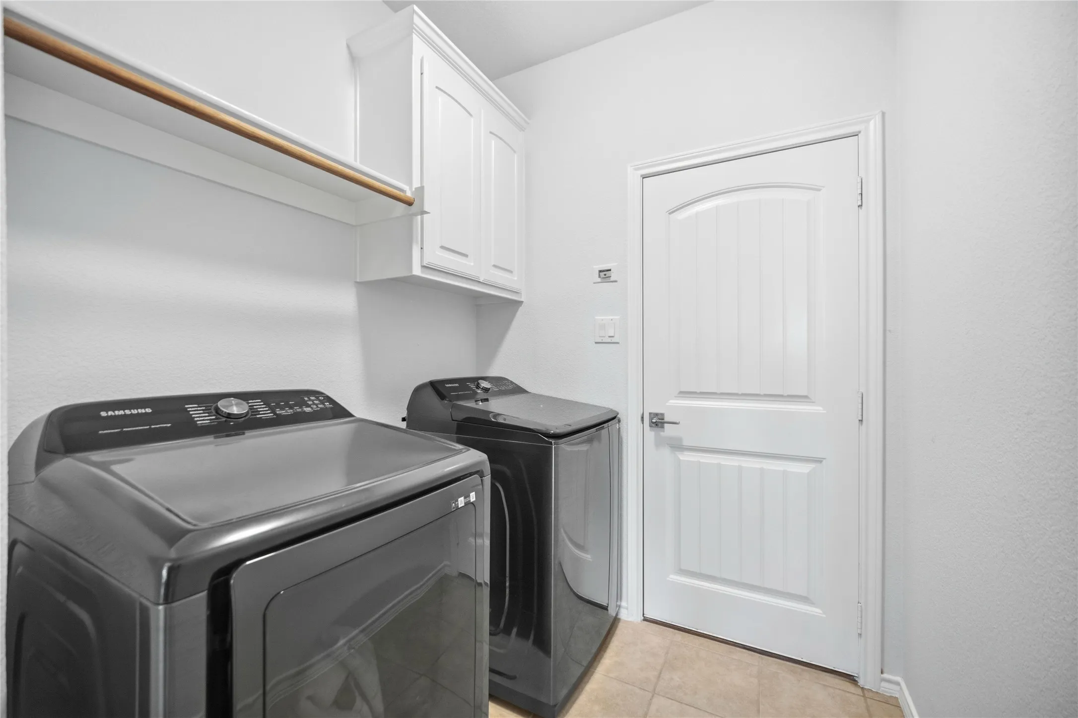 Dedicated laundry space featuring white wall-mounted cabinetry, an upper shelving unit with a hanging rod, a paneled door, and neutral-toned tile flooring