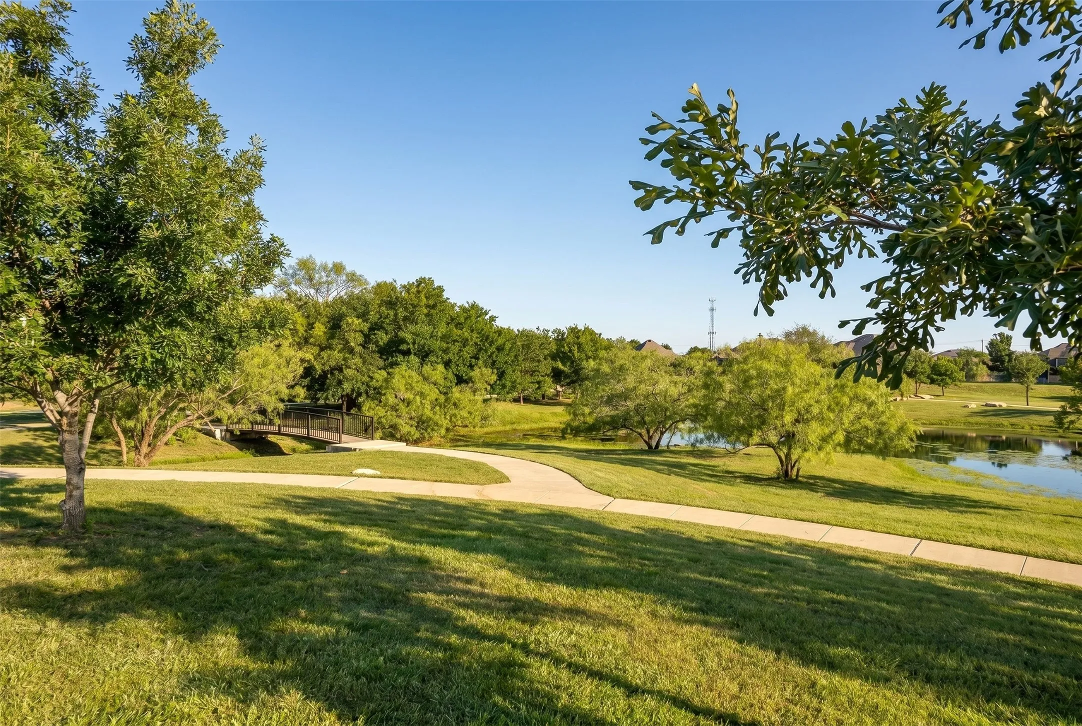 Landscaped grounds featuring a paved walking path, mature trees, a tranquil pond, and a pedestrian bridge