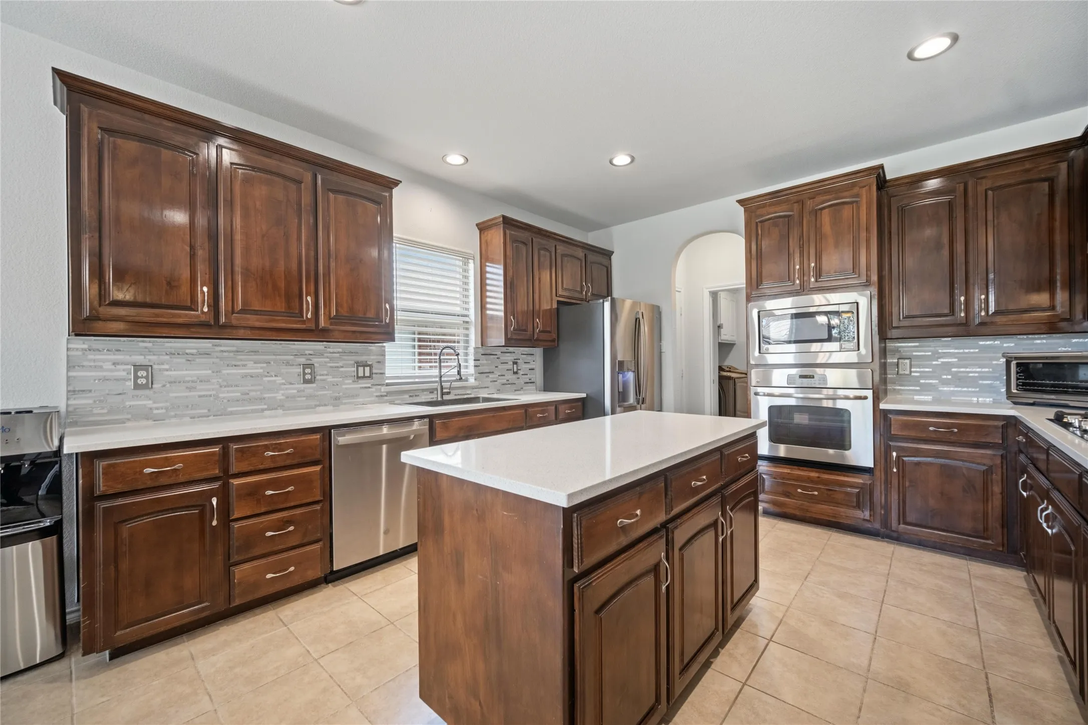 Kitchen featuring warm wood cabinetry, stainless steel appliances including a built-in oven and microwave, light-toned countertops, and a tiled backsplash