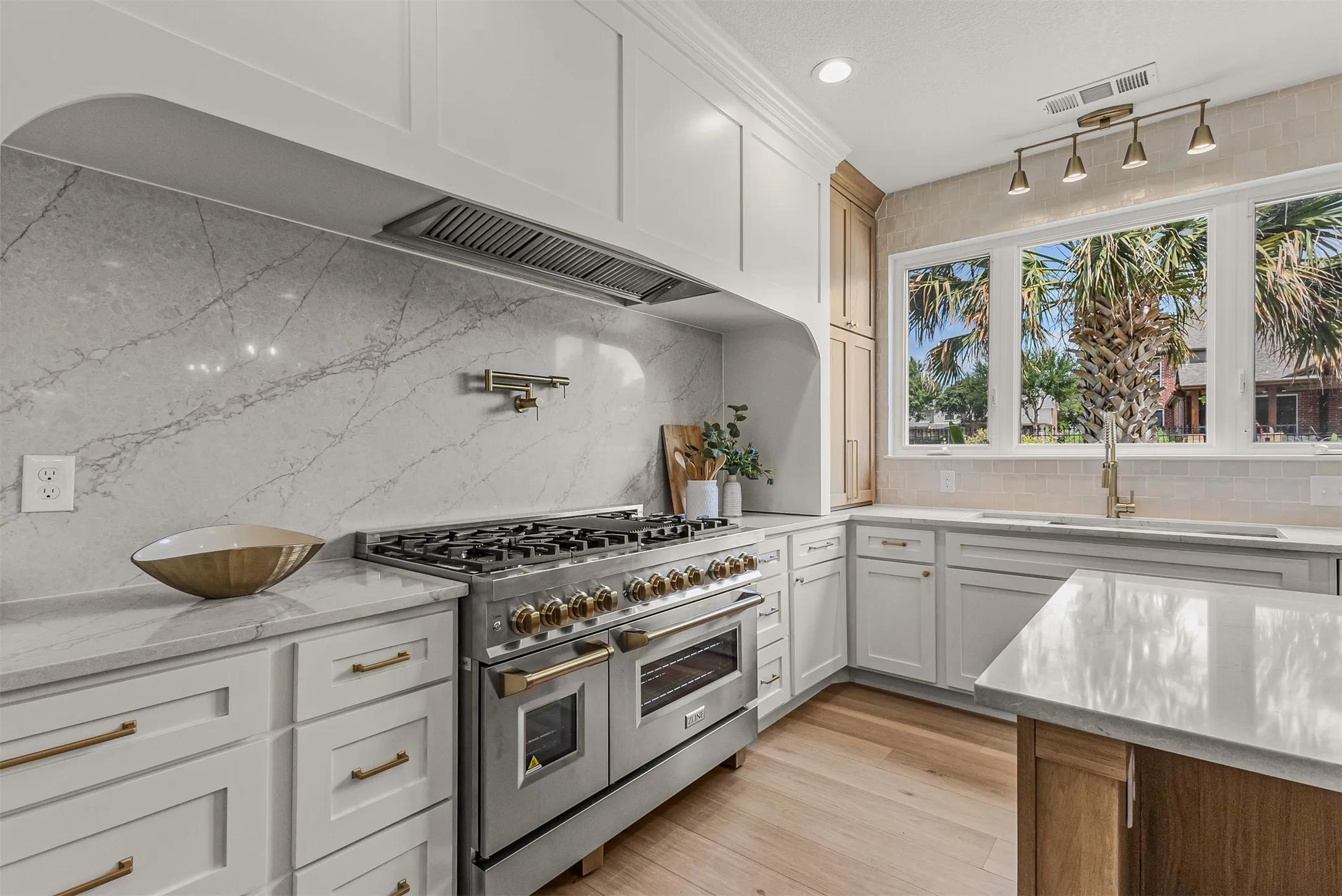 Gourmet kitchen featuring a professional-grade stainless steel range, custom range hood, slab backsplash, light wood-finish flooring, and white perimeter cabinetry with gold-tone hardware