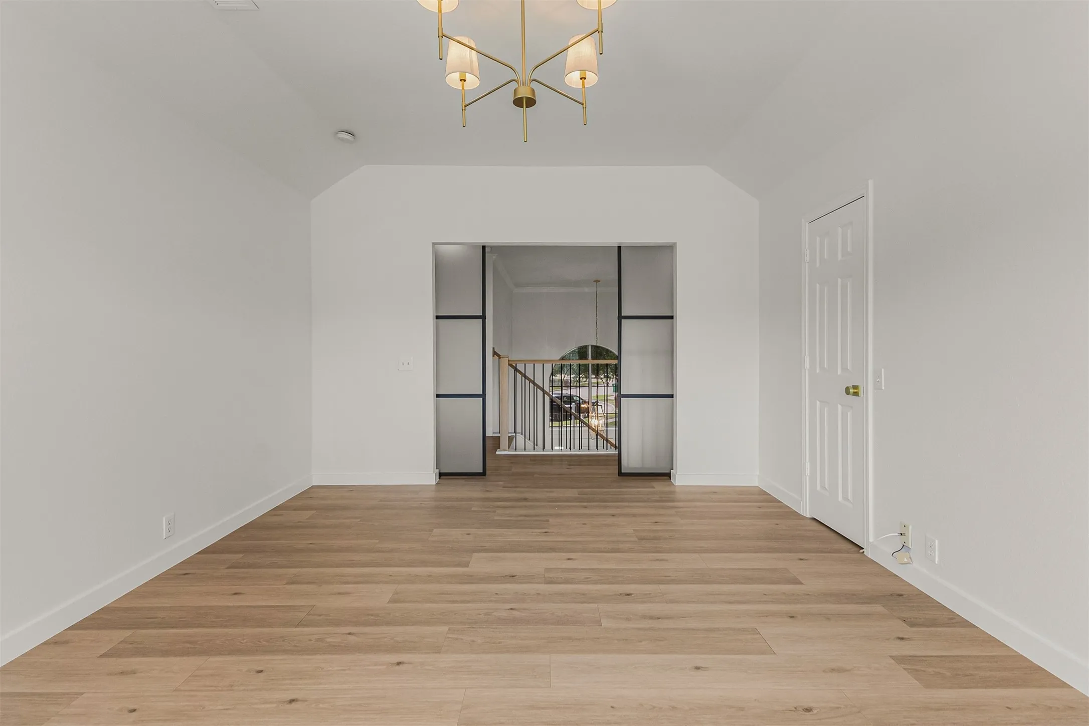 Spacious room featuring light wood-finish flooring, a contemporary brass chandelier, a paneled door, and a recessed entryway with a frosted glass and black metal frame