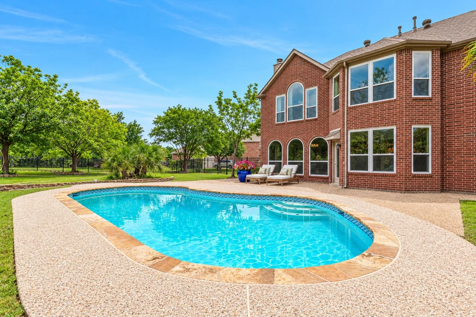 Brick exterior with an oval-shaped pool, exposed aggregate patio, and mature trees
