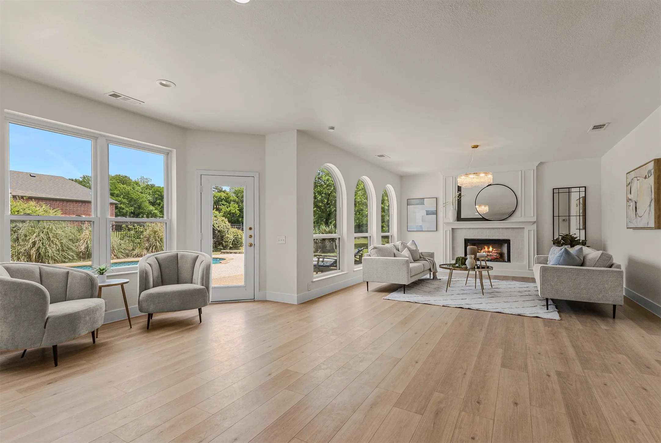 Expansive living area featuring light wood-finish flooring, a white fireplace with ornate trim, multiple windows with arched details, and an exterior door leading to a visible pool area
