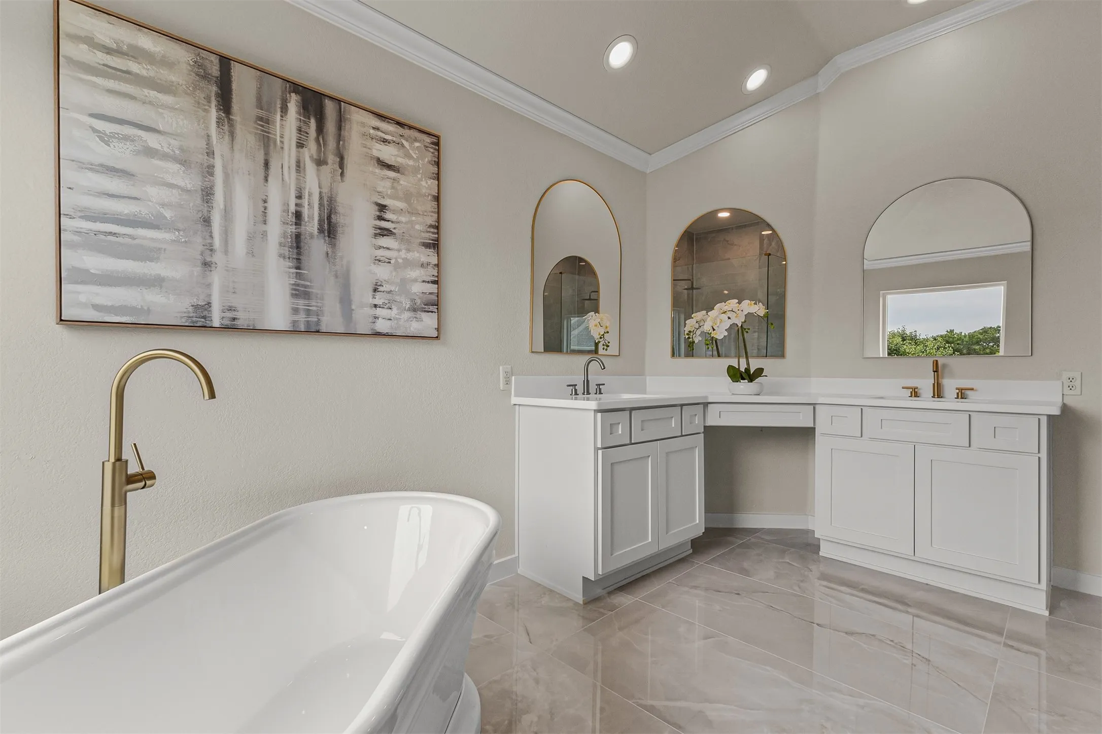Freestanding soaking tub with a floor-mounted brushed gold faucet, dual vanities with white cabinetry and countertops, arched mirrors, and large format polished tile flooring