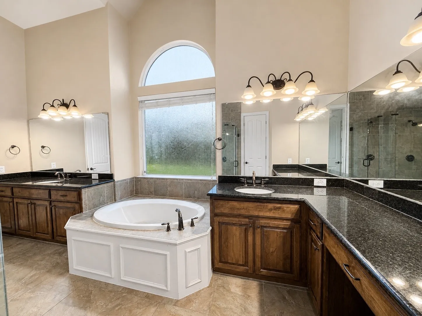 Expansive bathroom featuring a corner soaking tub, dual wood-finish vanities with dark stone countertops, and a tiled shower enclosure visible in reflection