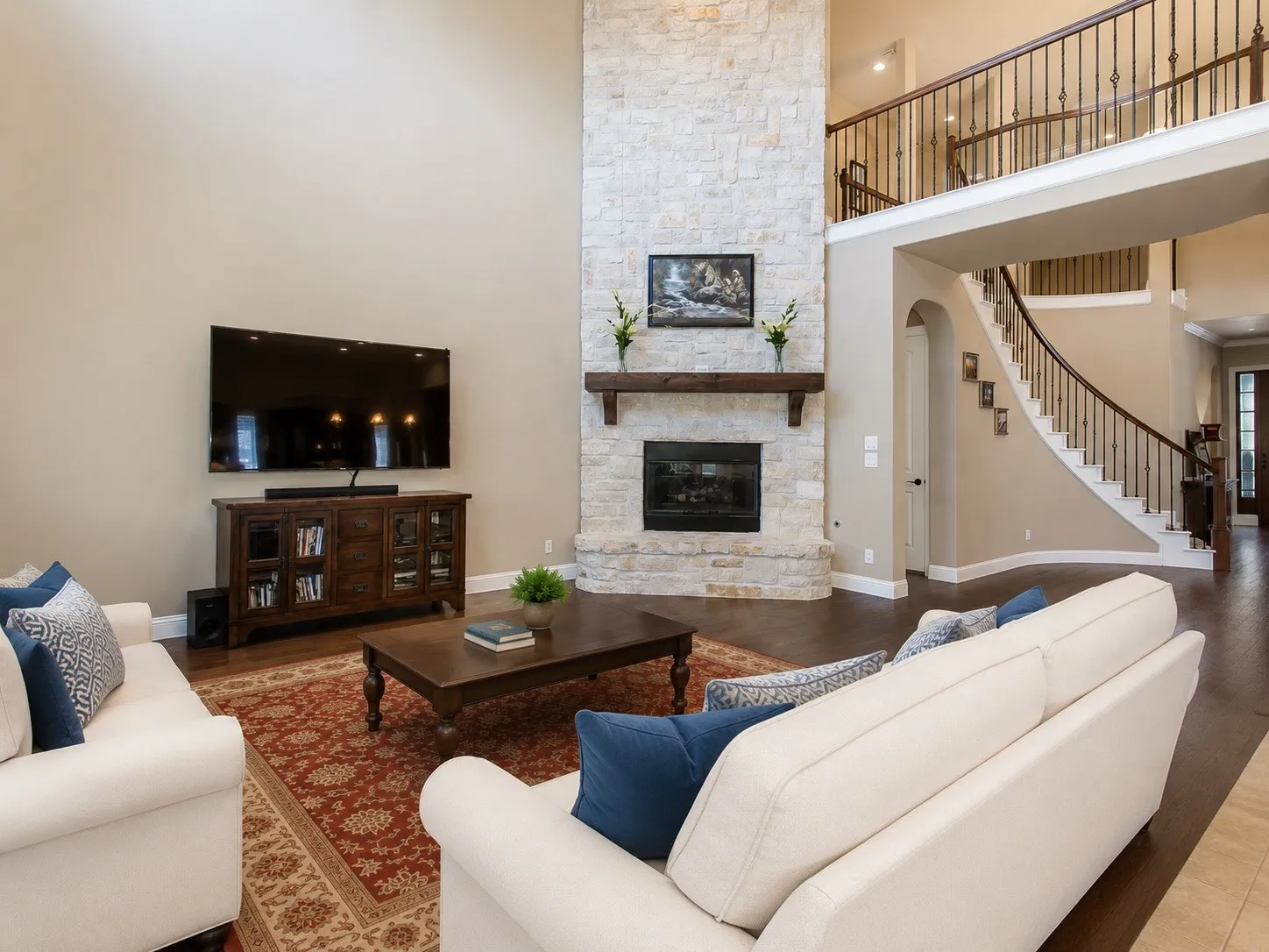 Two-story living area featuring a stone-clad fireplace with a wood mantle, rich wood-finish flooring, and a curved staircase with wrought iron balusters