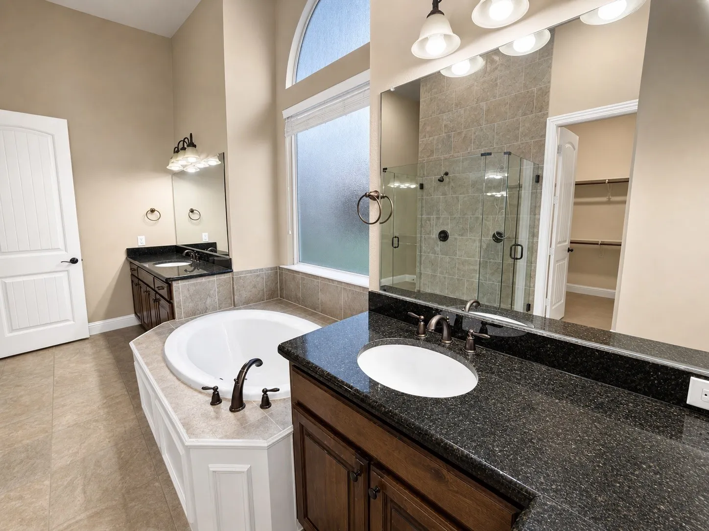 Bathroom featuring a built-in garden tub with white paneling, dual vanities with dark countertops and integrated sinks, a glass-enclosed shower with tiled walls, and a large arched window