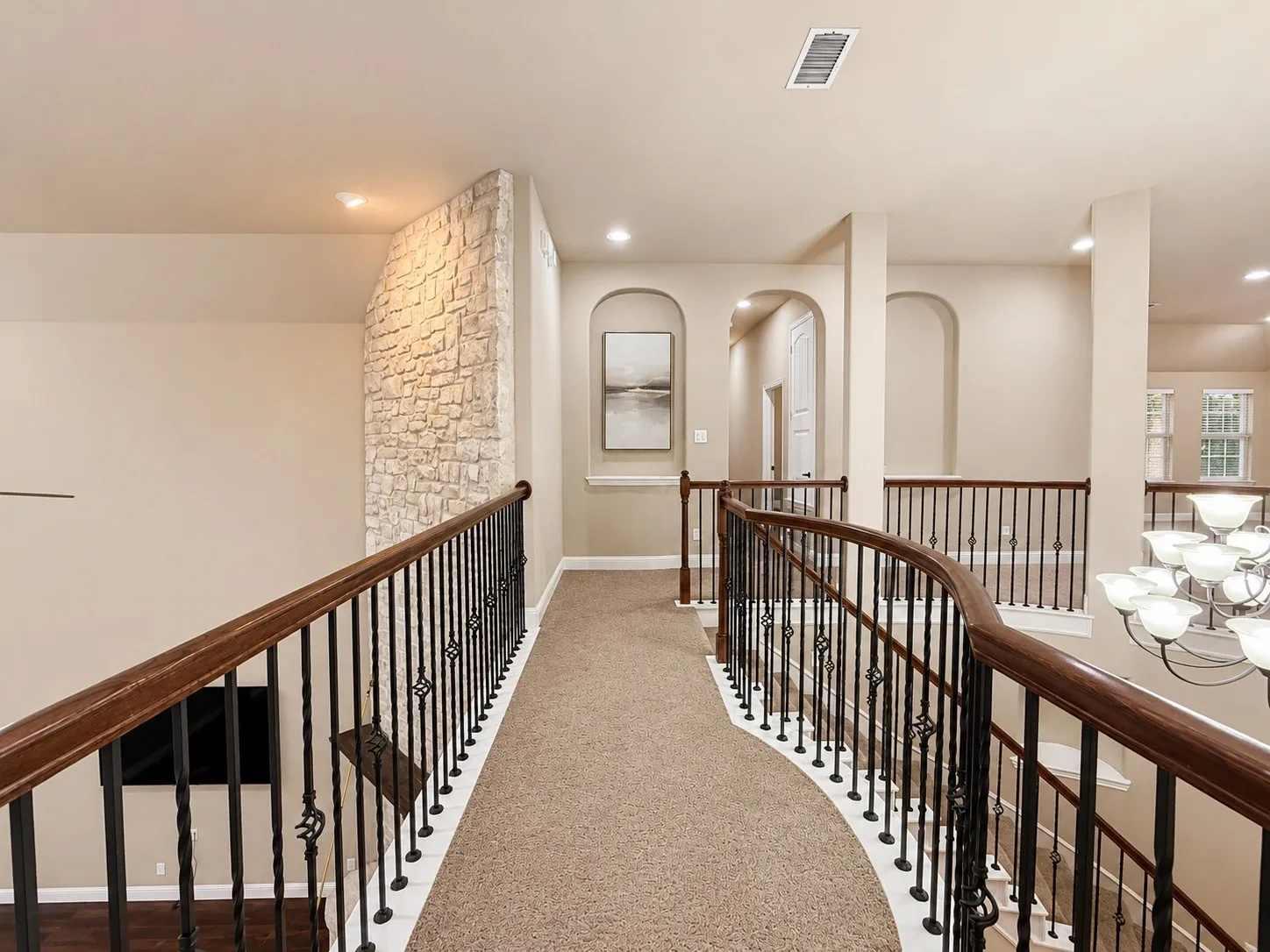 Second-story hallway featuring a curved wrought iron and wood-finish railing, plush neutral carpeting, stone accent wall, and recessed lighting