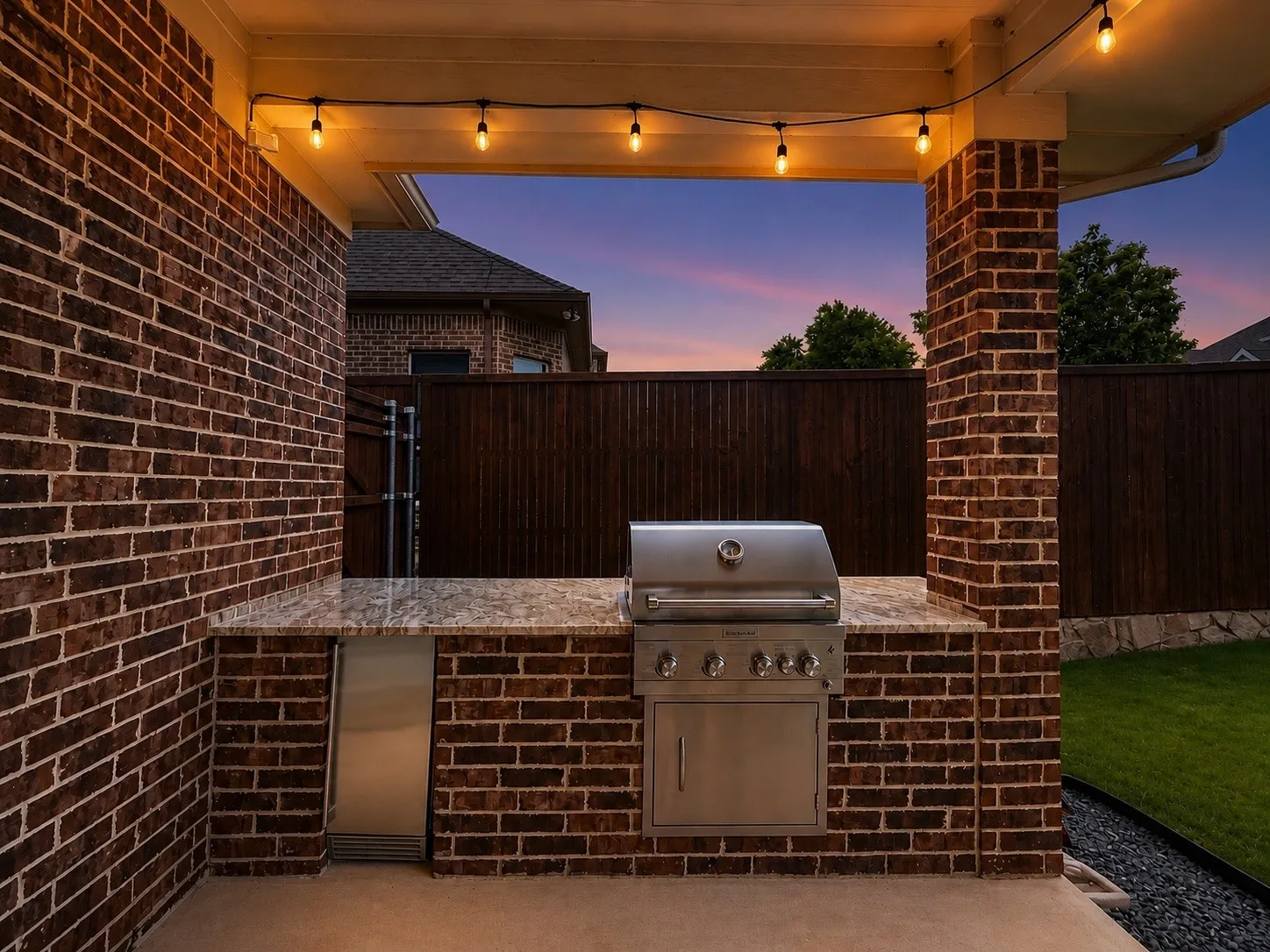 Outdoor kitchen featuring a built-in stainless steel grill, integrated stainless steel under-counter refrigerator, speckled countertop, brick base, and string lighting