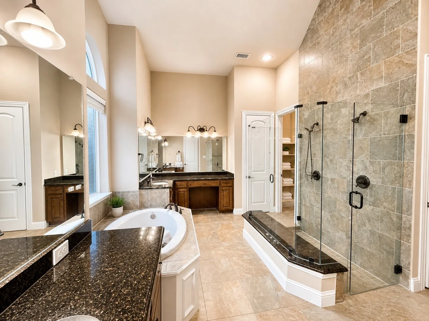 Spacious bathroom featuring a built-in tub, separate glass-enclosed shower with dual showerheads, dual vanities with dark stone countertops, and a tall arched window
