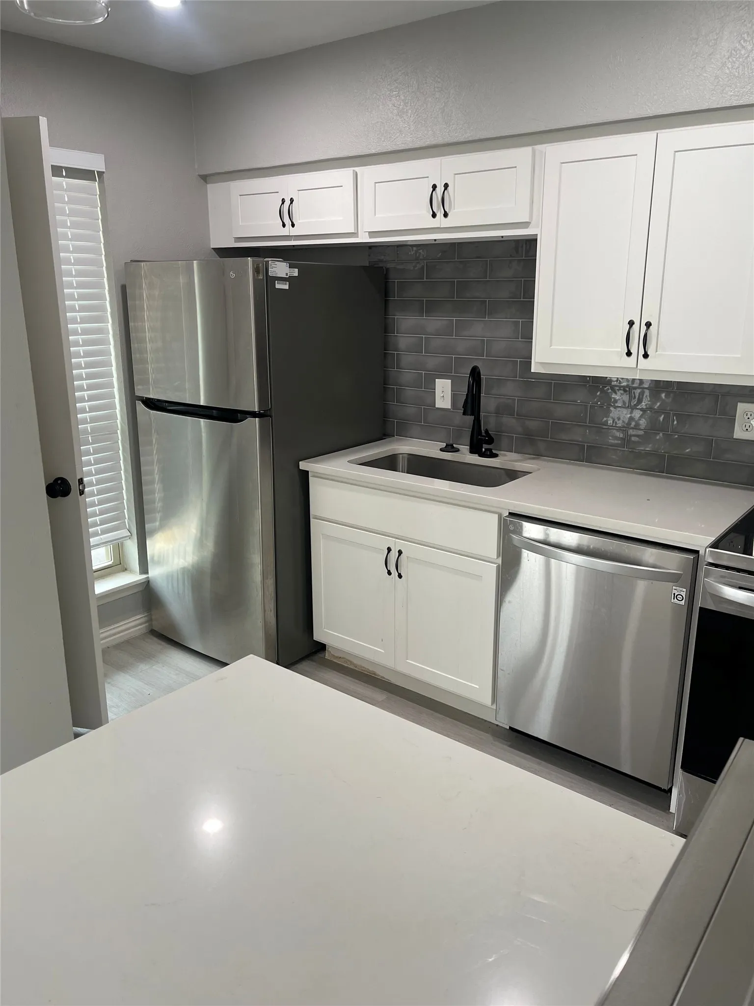Kitchen featuring white shaker-style cabinetry, a stainless steel sink with a matte black faucet, a dark gray subway tile backsplash, and light-toned countertops