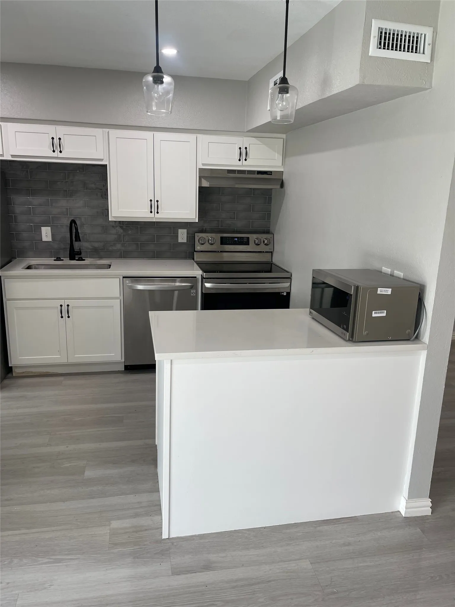 Modern kitchen featuring white shaker cabinetry, stainless steel appliances, a dark subway tile backsplash, light-toned wood-finish flooring, and pendant lighting