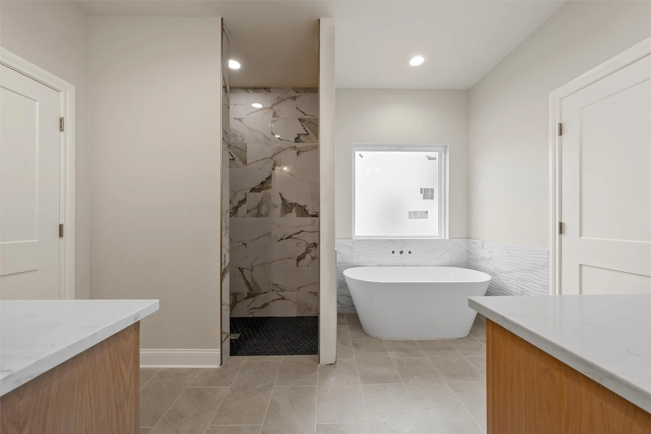 Spacious bath featuring a freestanding soaking tub, a walk-in shower with large format marble-look tile, natural wood-finish vanity bases with light countertops, and light gray floor tiling