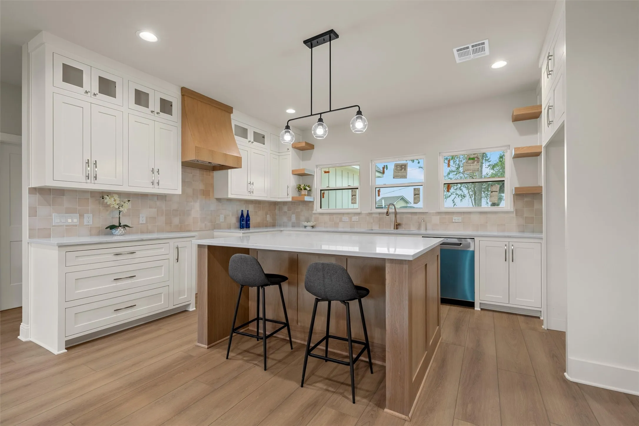 Open-concept kitchen featuring an expansive center island with seating, white shaker-style cabinetry, wood-finish flooring, a light wood range hood, and a light-toned tile backsplash