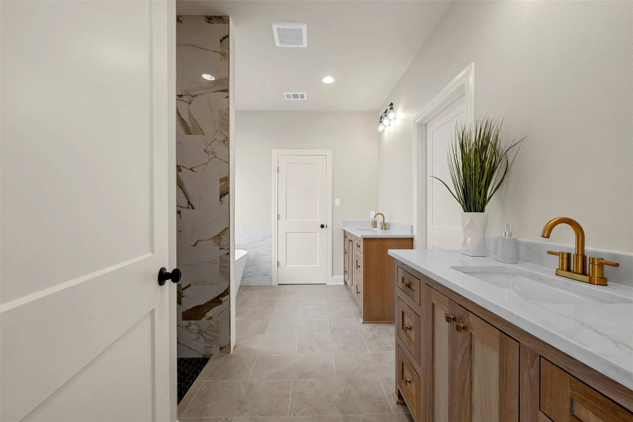 Bathroom featuring dual wood-finish vanities with white countertops, gold-finish fixtures, a freestanding bathtub, and a walk-in shower with natural stone-look tiling