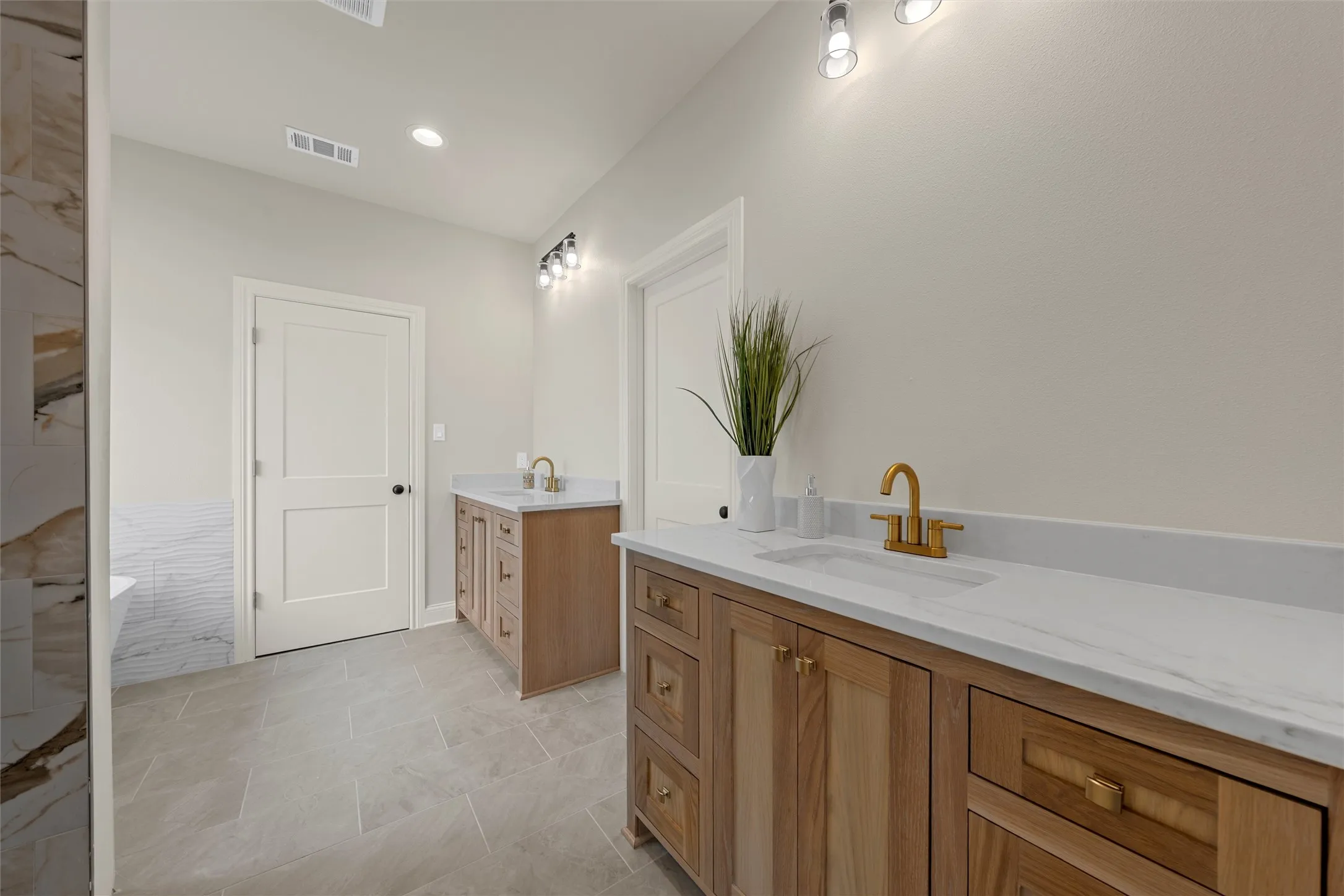 Dual wood-finish vanities with undermount sinks and gold-tone fixtures, complemented by a stone-look tile floor and recessed lighting