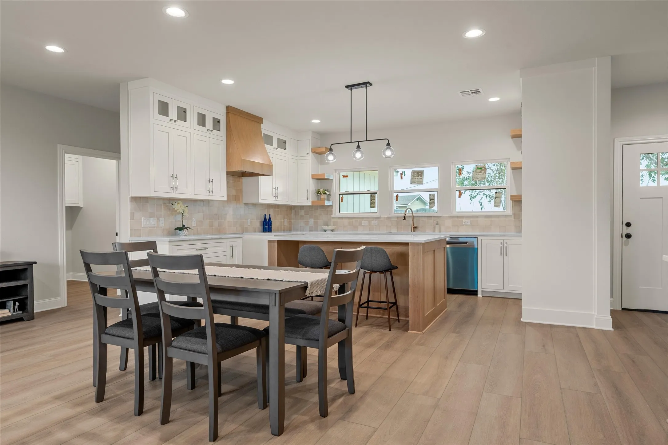 Bright kitchen and dining area featuring light wood-finish flooring, white cabinetry, a wood range hood, a tiled backsplash, and a central island with bar seating