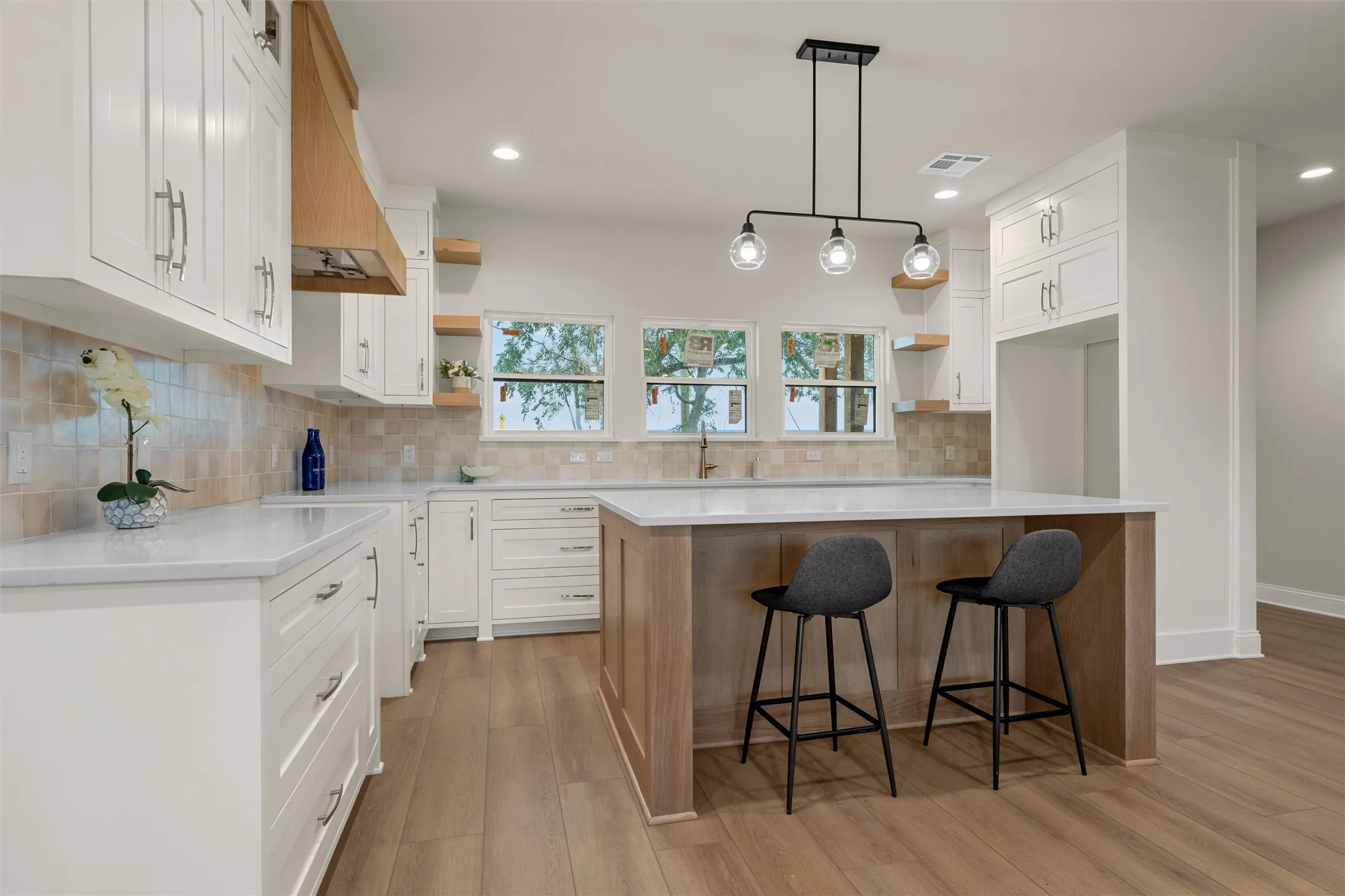 Bright kitchen featuring white shaker cabinetry, light wood-finish flooring, a central island with countertop seating, a light wood-finish range hood, and a geometric pendant light fixture