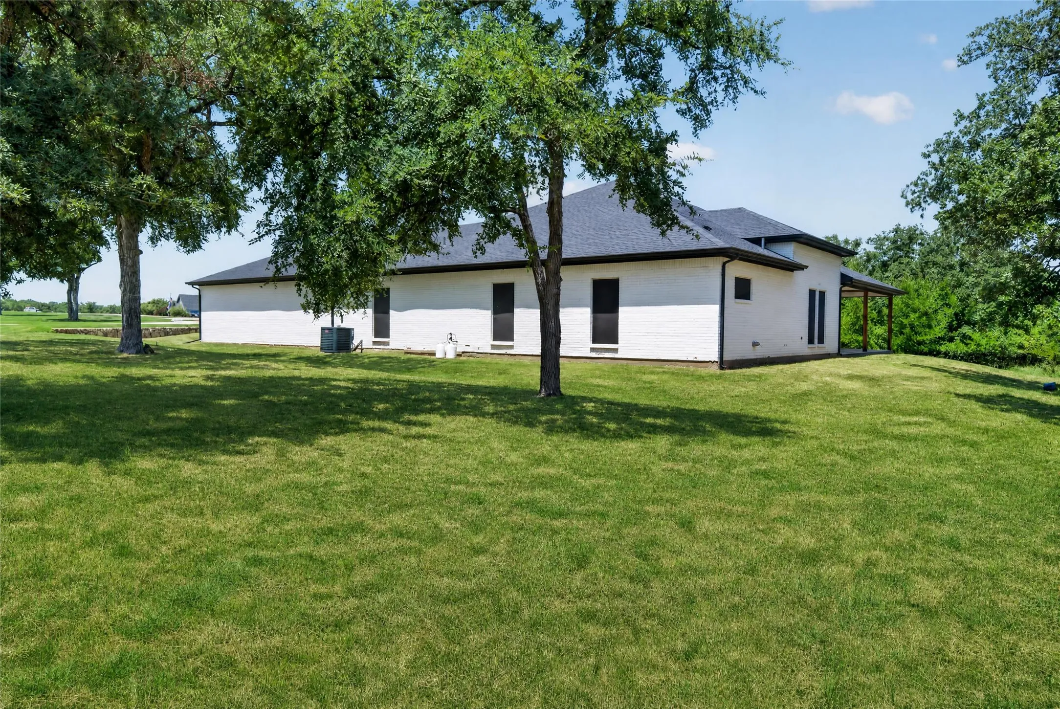 Spacious lawn with mature trees surrounding a white brick exterior featuring black-trimmed windows and a dark roof