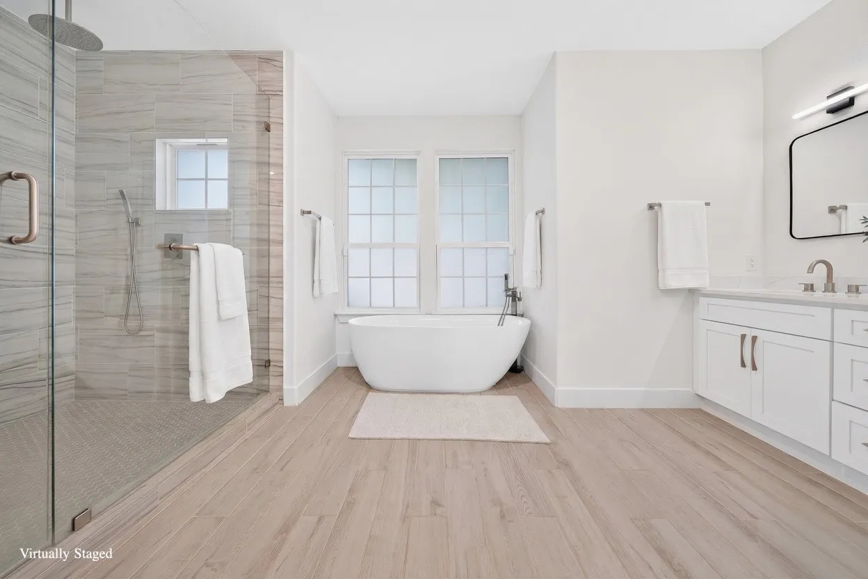 Spacious bathroom featuring a freestanding soaking tub, a frameless glass shower with rain head and handheld fixtures, a dual-sink vanity with white cabinetry, light wood-finish flooring, and frosted windows