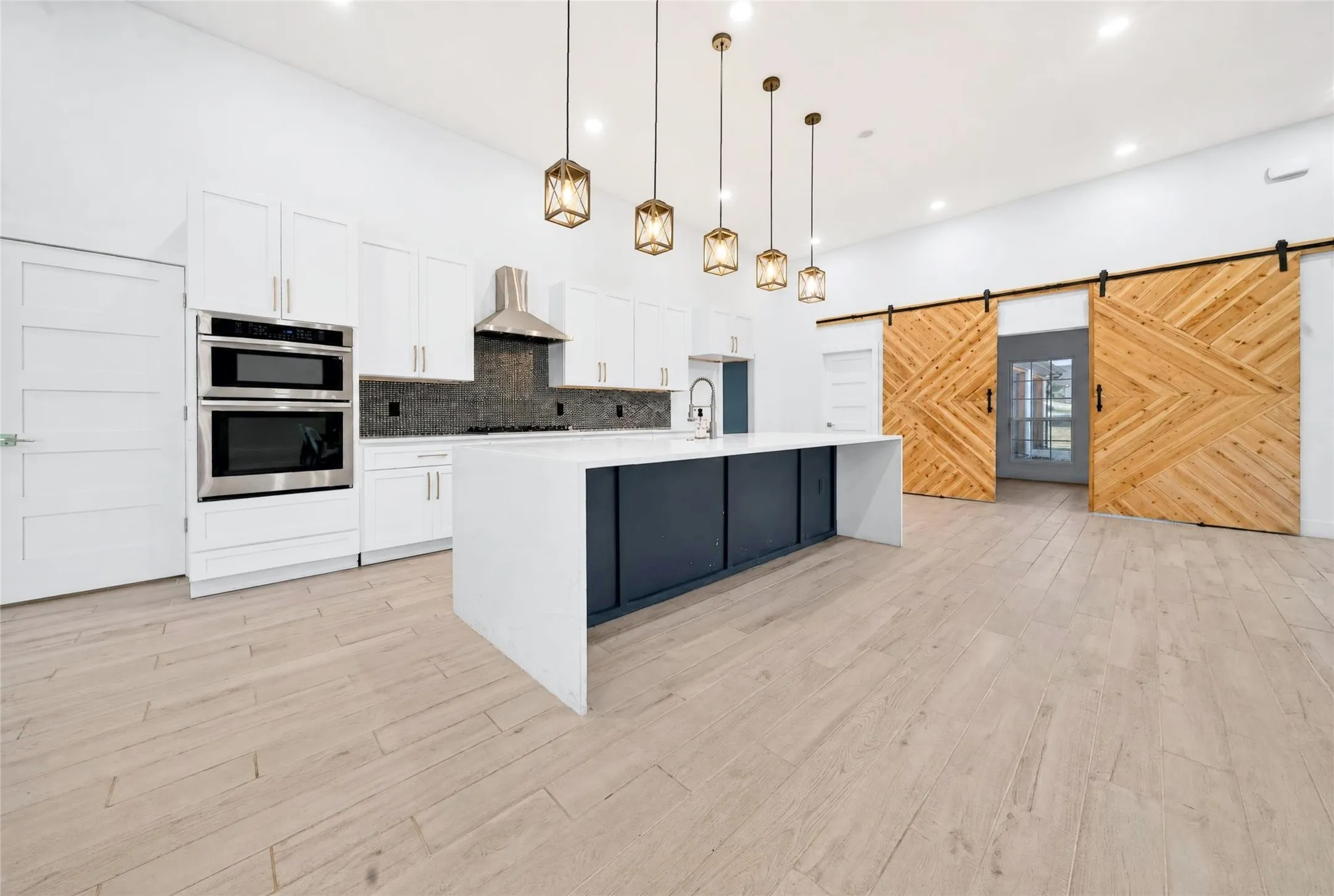 Modern kitchen featuring white shaker cabinetry, stainless steel appliances, a dark backsplash, and an oversized island with a waterfall countertop