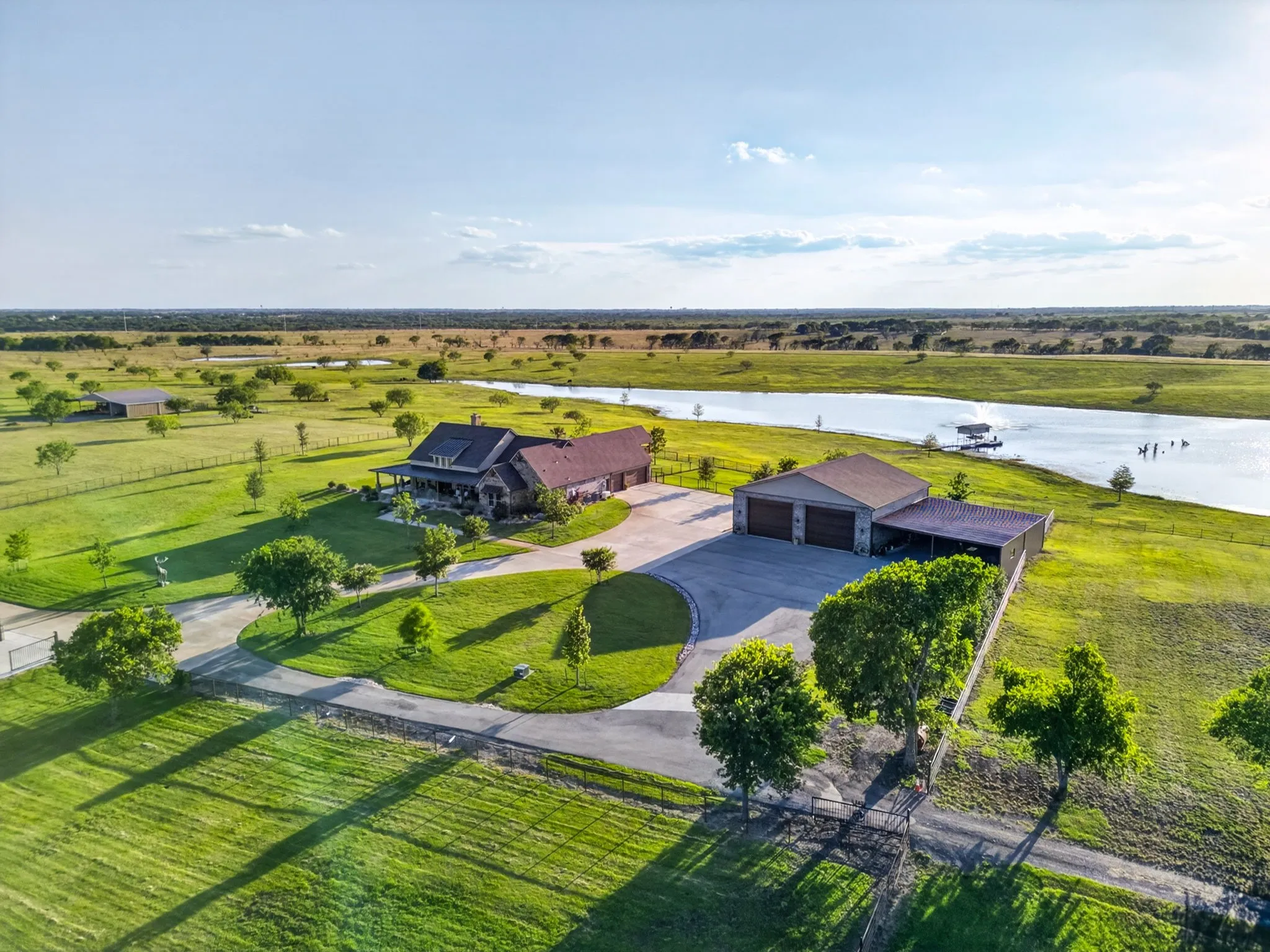 Expansive property featuring a large body of water, a main residence with a covered porch, a detached garage structure with multiple bays and an attached carport, and a prominent paved driveway