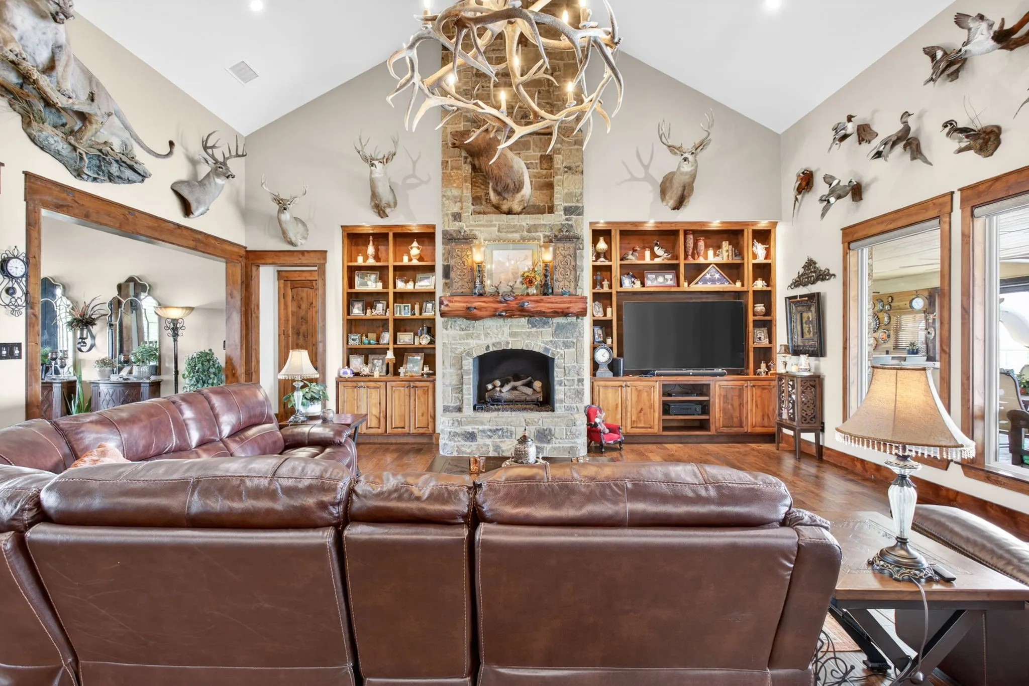 Vaulted ceiling family room featuring a stone fireplace with a wood mantle, built-in cabinetry, wood-finish flooring, and large windows with wood trim