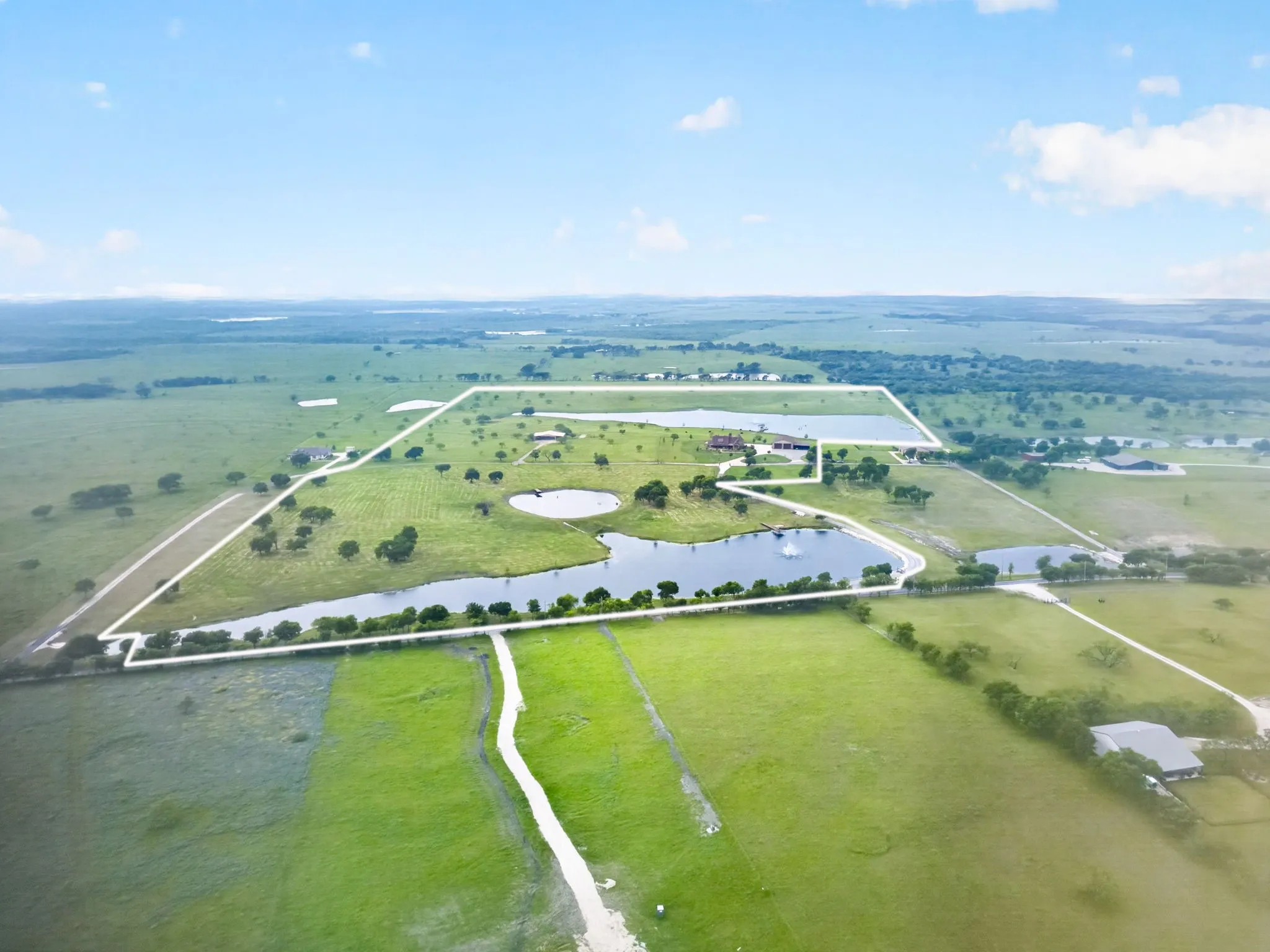 Expansive aerial depiction of a large land parcel featuring multiple water bodies, abundant green acreage, perimeter fencing, a primary residence with an attached garage, and additional outbuildings