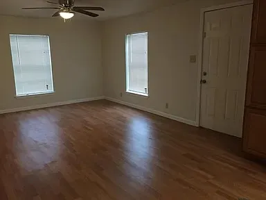 Spacious room featuring wood-finish flooring, two windows with blinds, a ceiling fan with integrated lighting, a paneled door, and built-in cabinetry