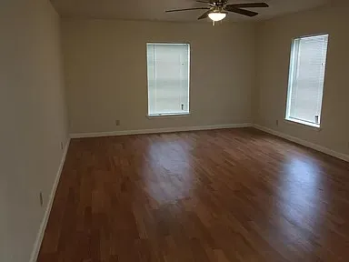 Spacious room featuring wood-finish flooring, two windows with blinds, a ceiling fan with integrated lighting, and neutral wall tones