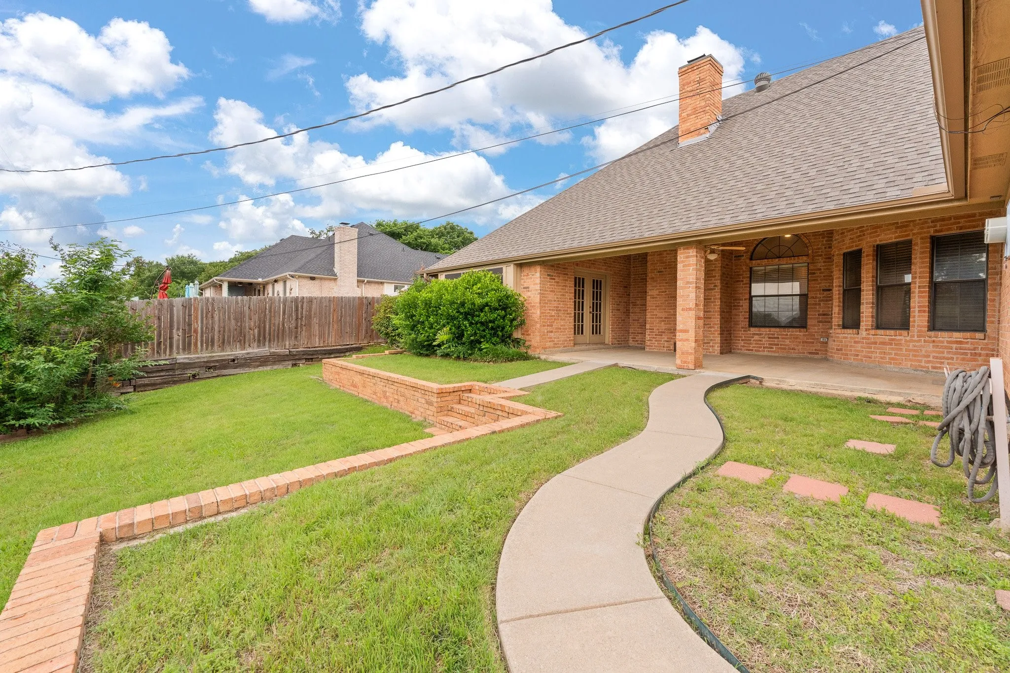 Expansive backyard featuring a winding concrete pathway, brick-retained planters, and a covered patio​​‌​​​​‌​​‌‌​‌‌​​​‌‌​‌​‌​‌​​​‌​​ area