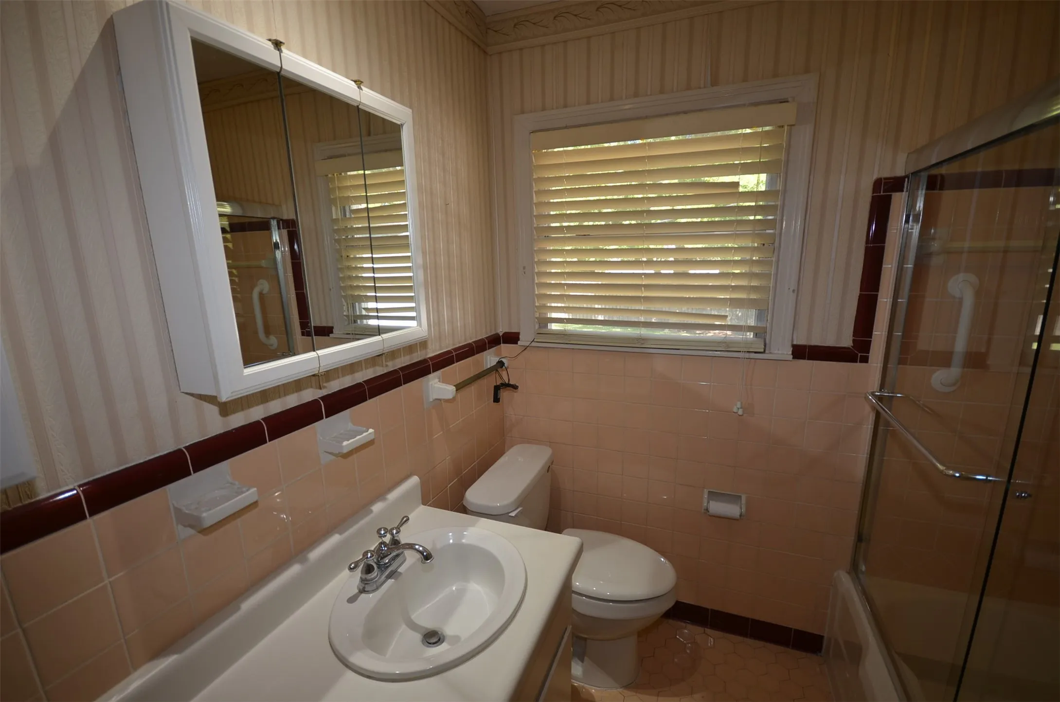 Bathroom featuring a vanity with an integrated sink, a mirrored medicine cabinet, a glass-enclosed shower, a window with blinds, and tiled walls