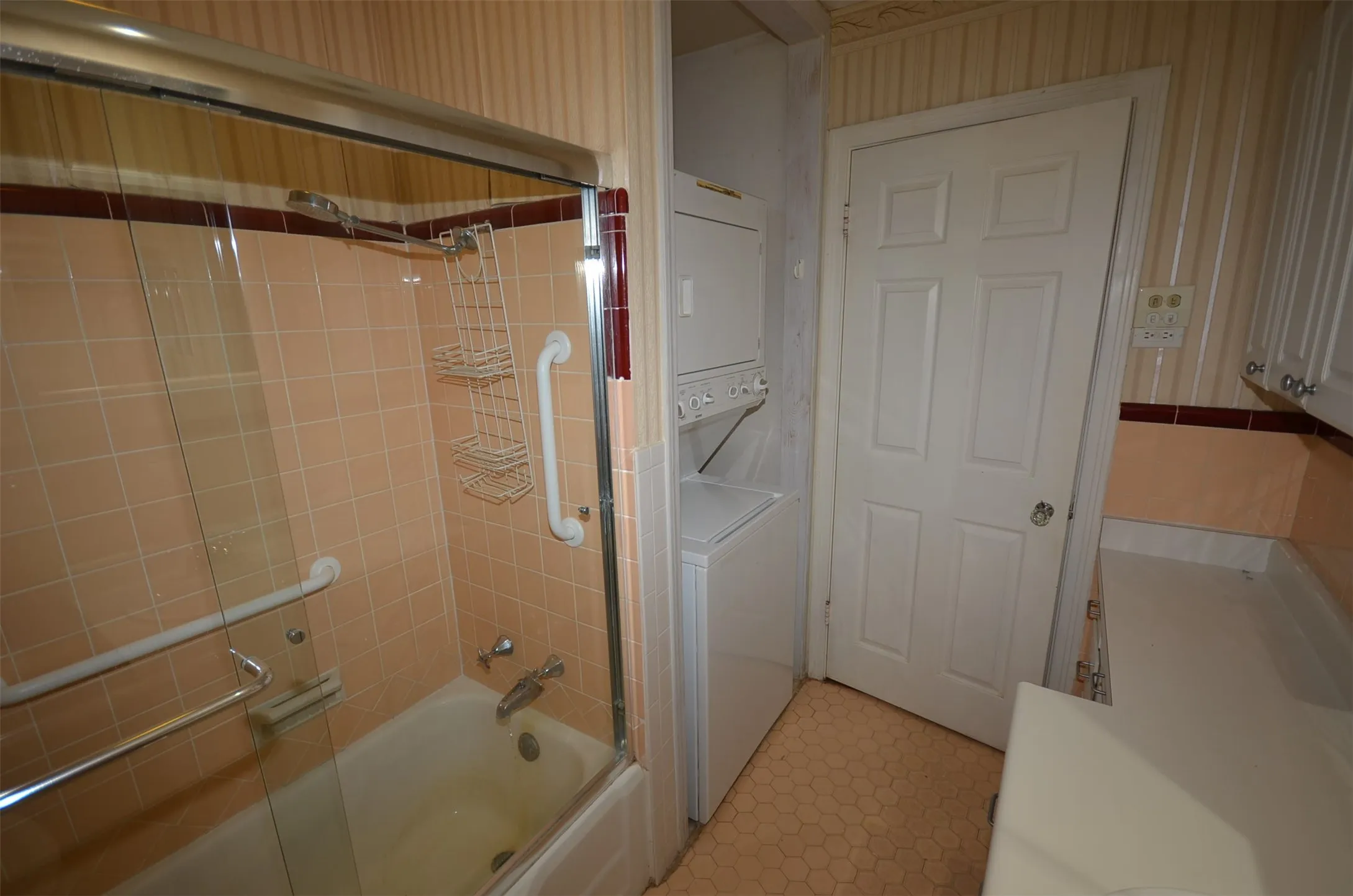 Bathroom featuring a tub/shower combination with glass enclosure, stackable washer and dryer, hexagon-patterned floor tiling, and a white countertop with cabinetry