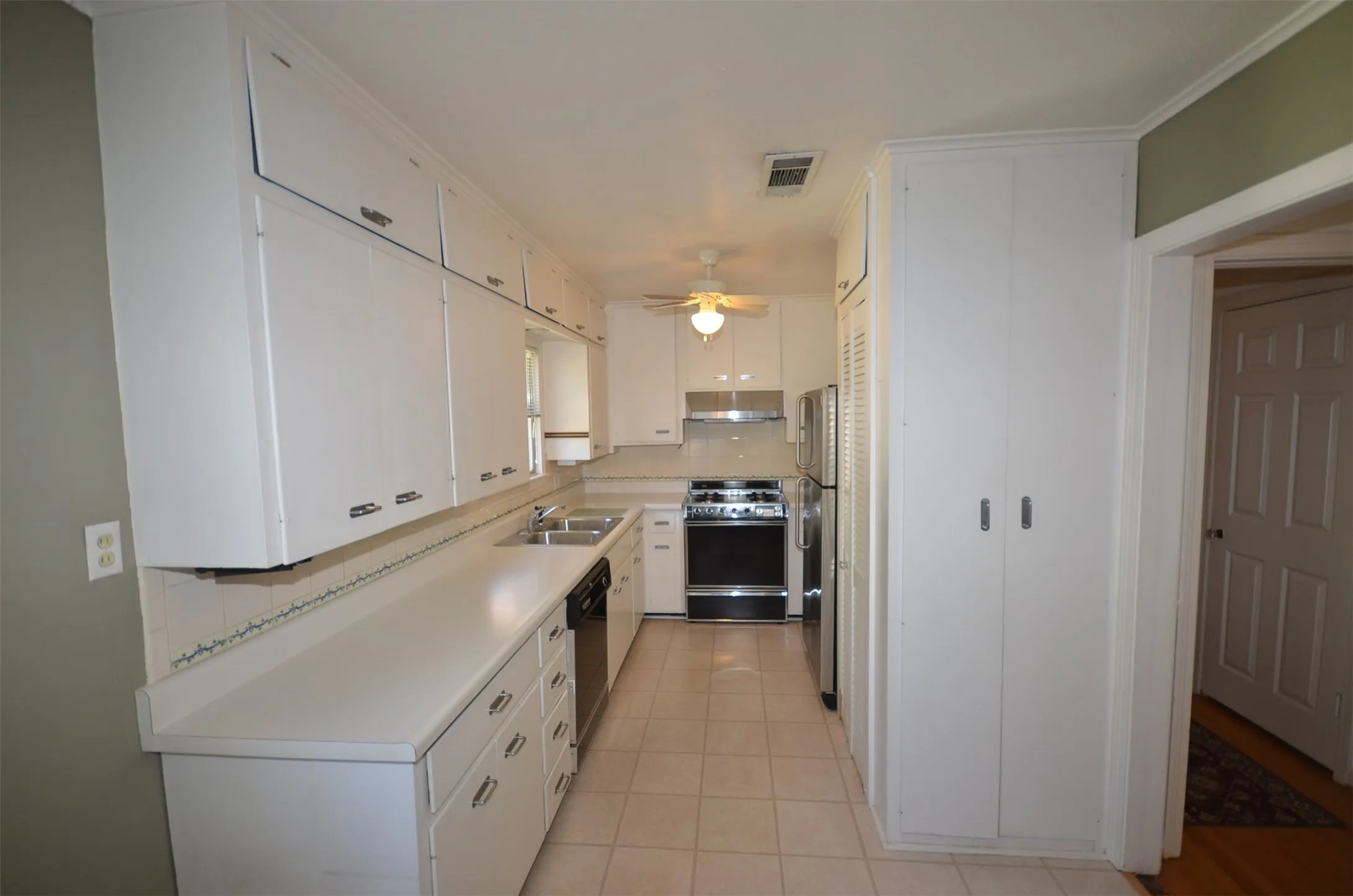 Galley kitchen featuring extensive white cabinetry, light-toned countertops, a double-basin stainless steel sink, tile flooring, and integrated appliances including a range and refrigerator