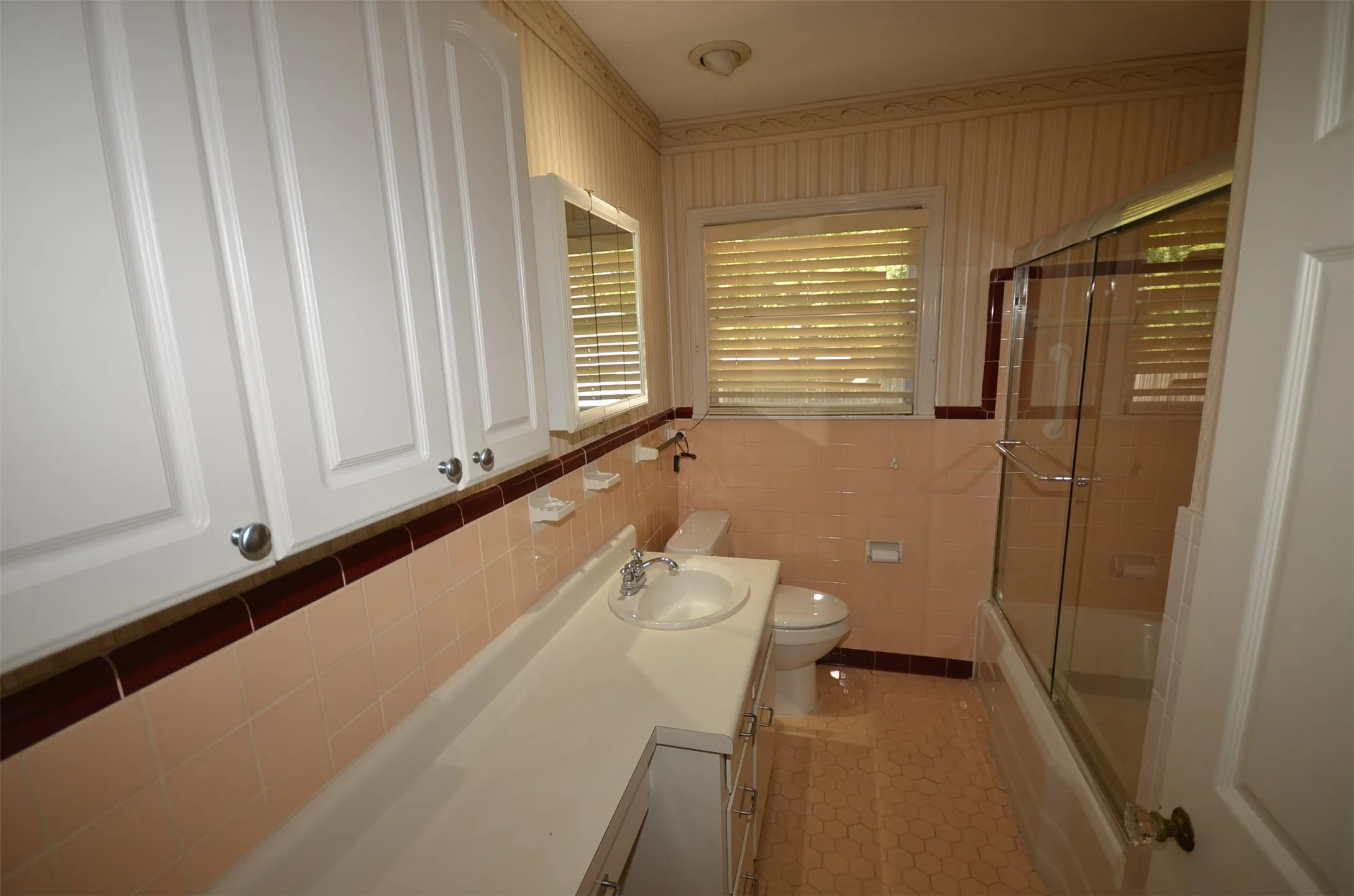 Bathroom featuring a vanity with an integrated sink, wall-mounted cabinetry, an enclosed shower-tub combination, and a window with horizontal blinds