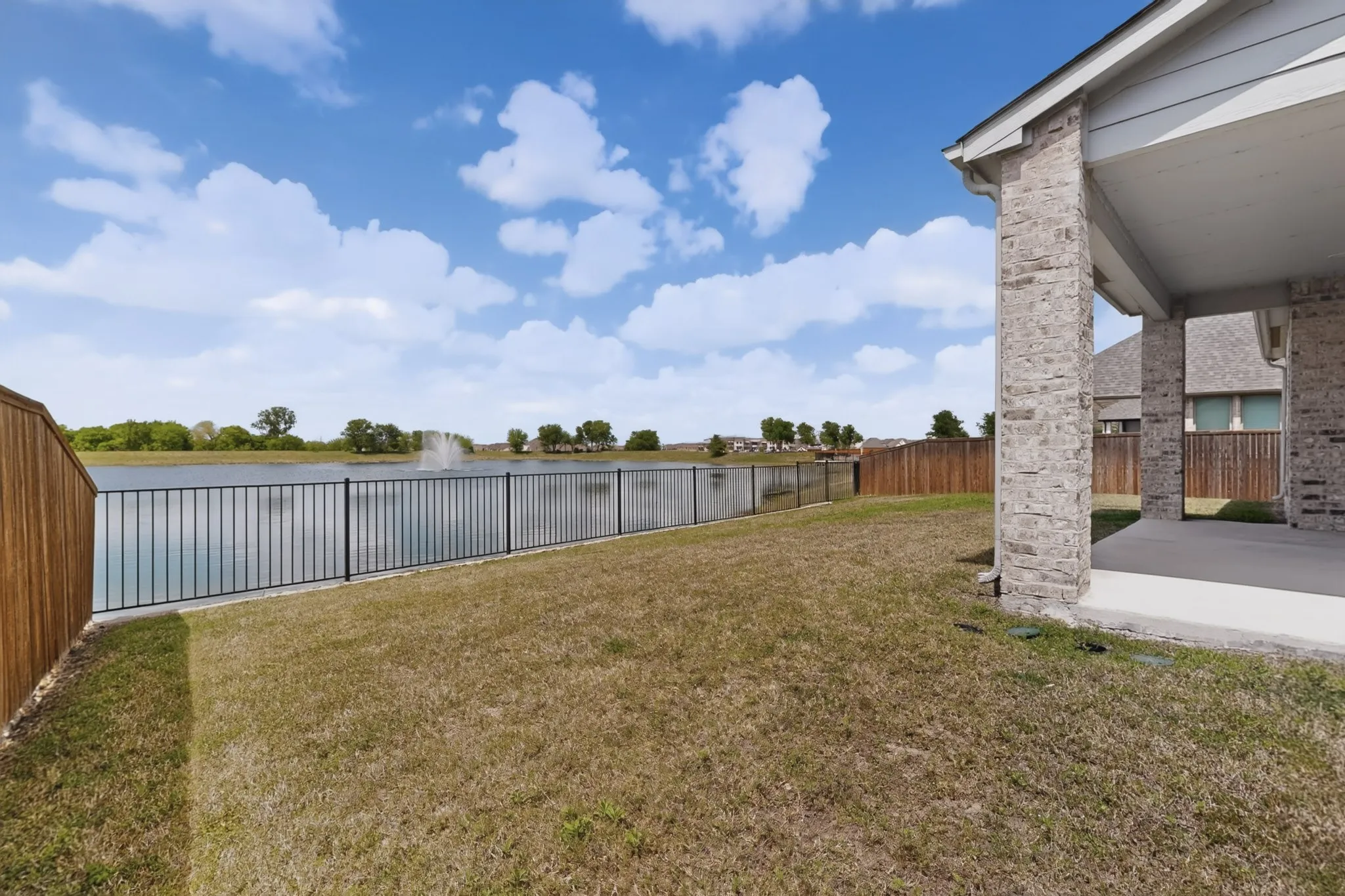 Backyard featuring a covered patio with brick columns, a metal fence, a wooden privacy fence, a lake with a fountain, and trees on the horizon