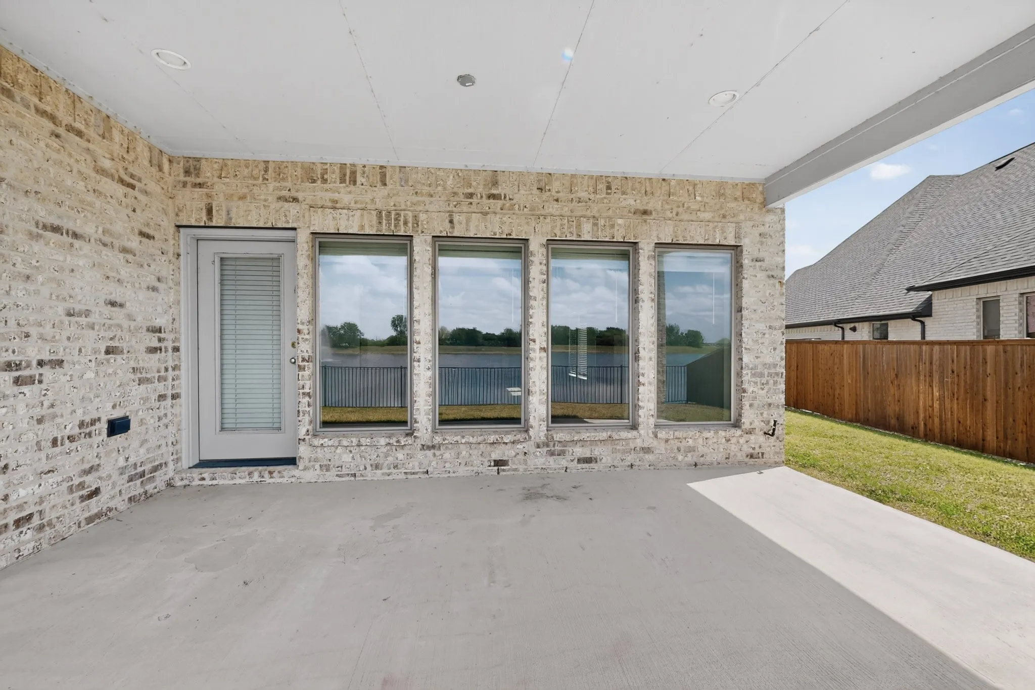 Covered concrete patio featuring recessed lighting, a brick exterior wall, and a door with integrated blinds, complemented by multiple large windows overlooking a body of water