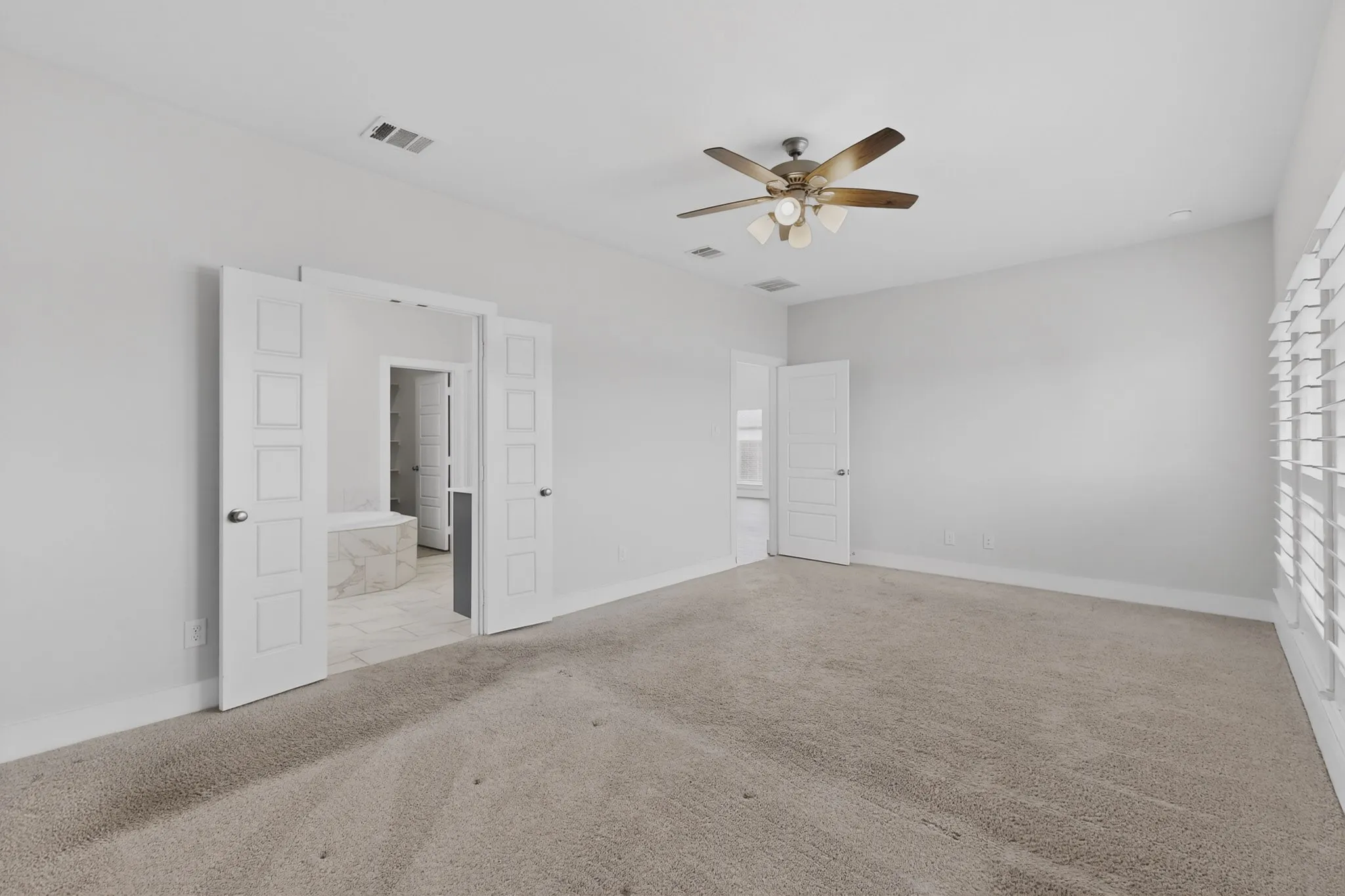 Spacious room with plush carpeting, ceiling fan with integrated lighting, and multiple windows featuring plantation shutters