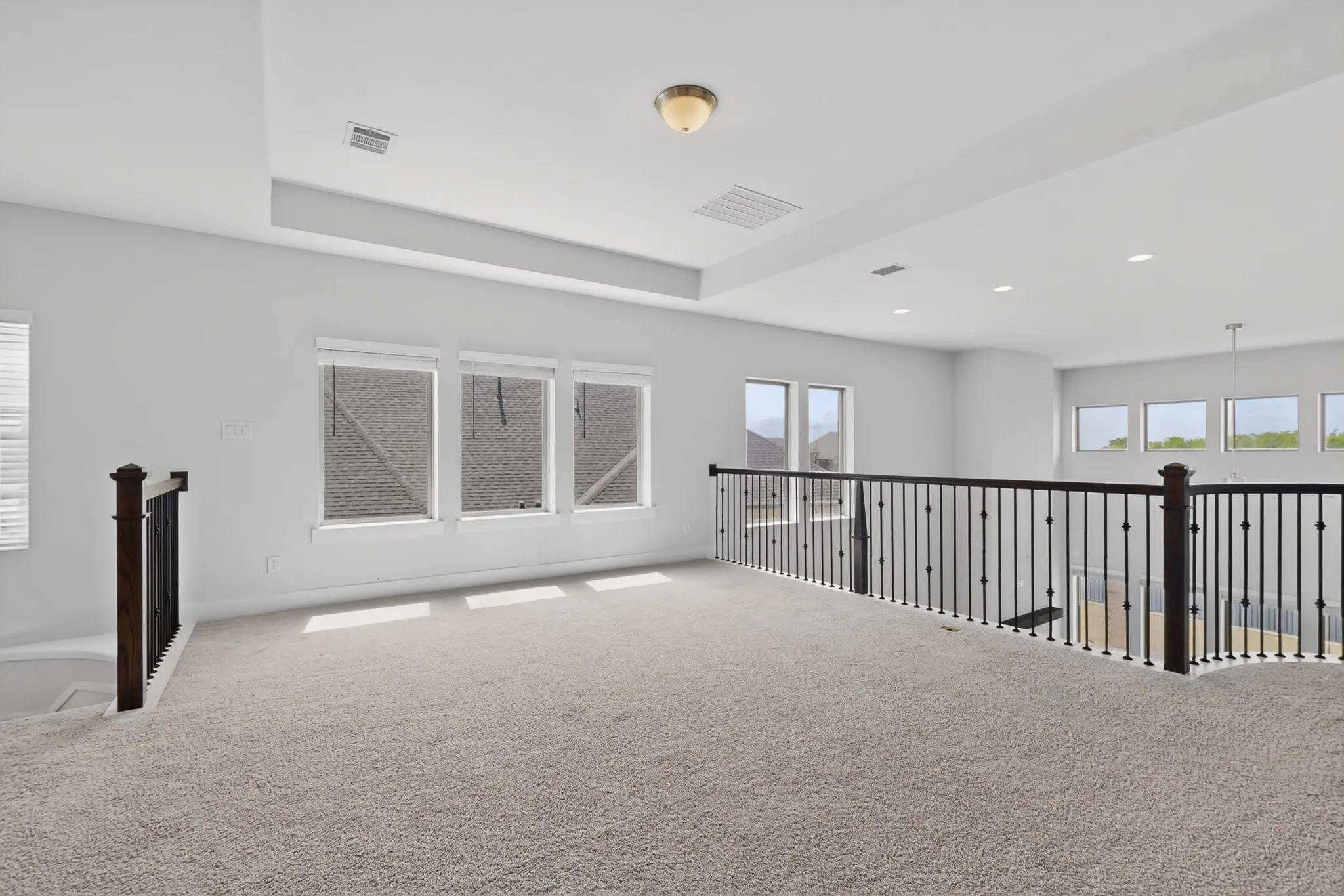 Spacious carpeted loft area featuring a tray ceiling, recessed lighting, multiple windows with blinds, and decorative wrought iron railing with wood newel posts