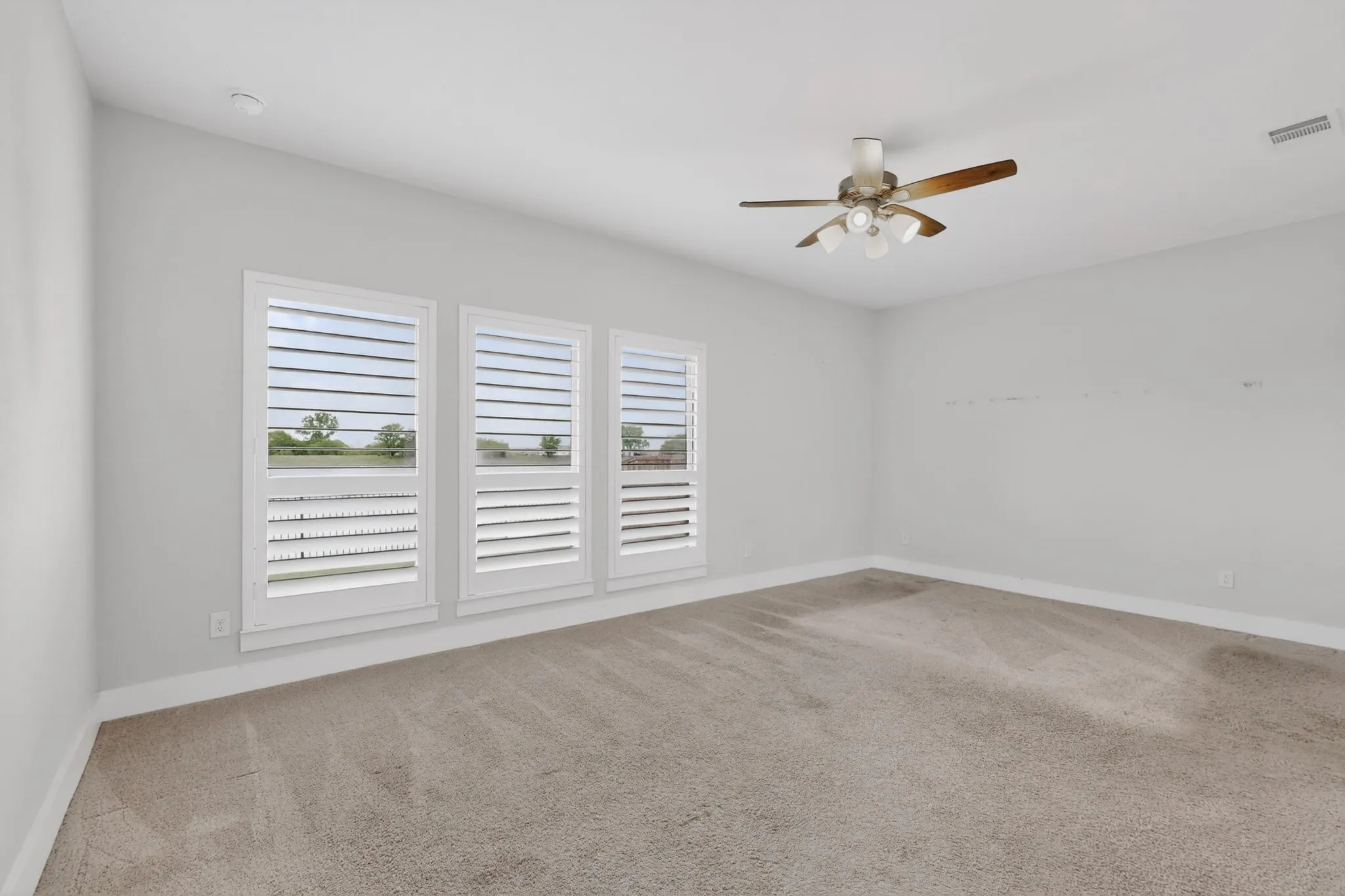 Spacious room featuring three windows with white plantation shutters, a ceiling fan with integrated lighting, and neutral-toned carpeting