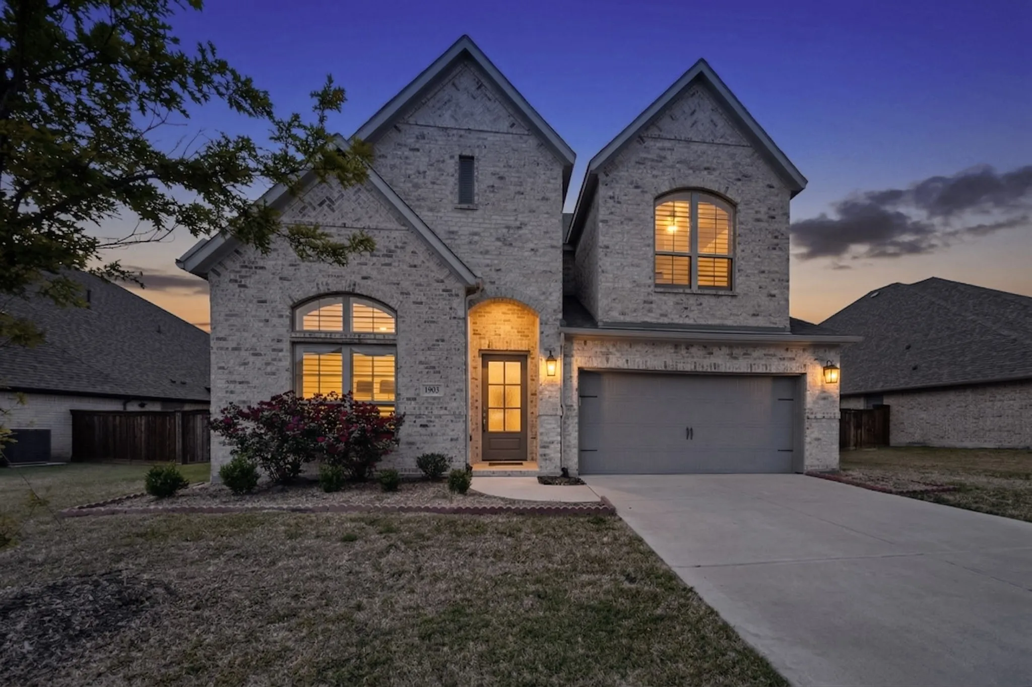 Two-story brick exterior featuring gabled roofs, arched windows with shutters, a covered entryway, an attached garage, and landscaped front yard