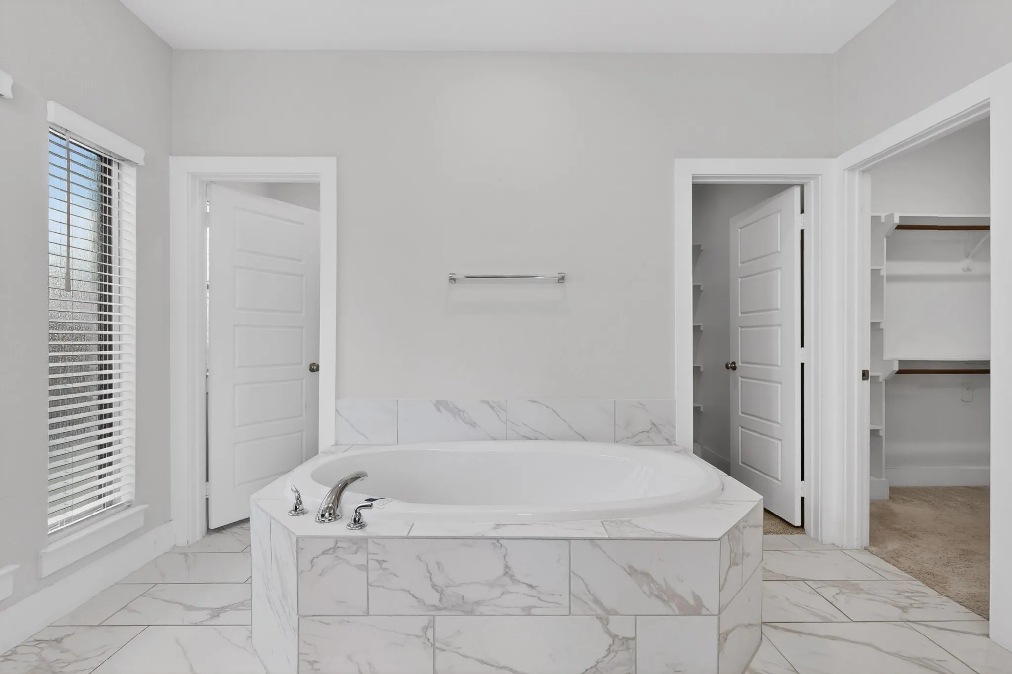 Soaking tub with a tiled surround, chrome fixtures, and a large window with blinds