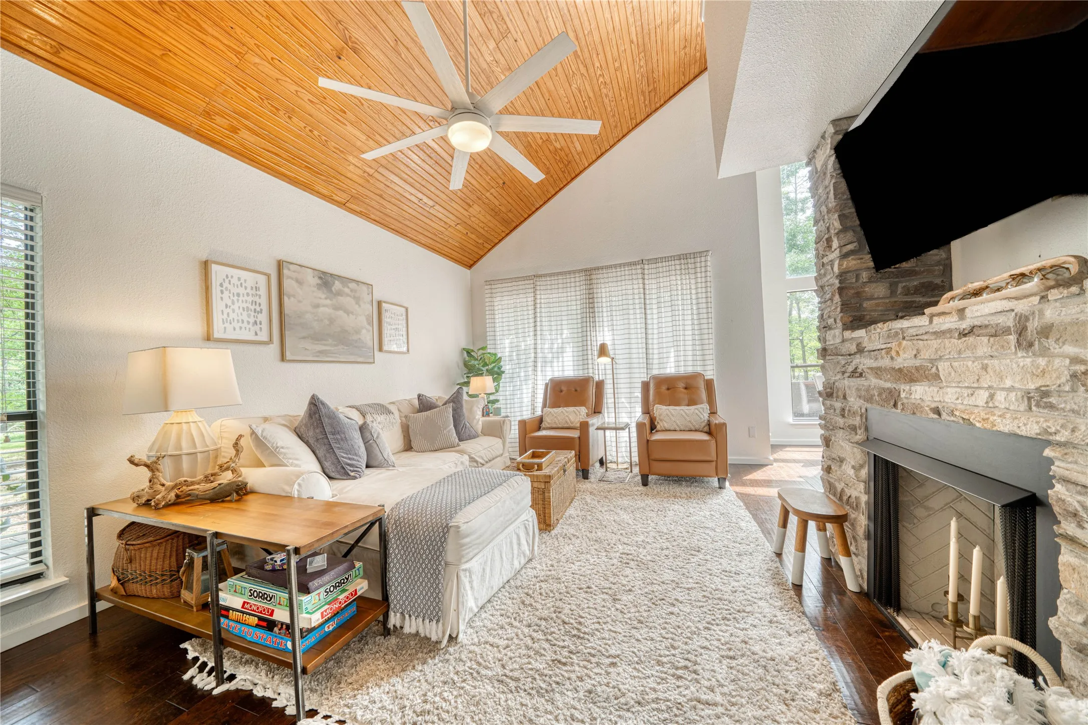 Living area with a vaulted wood-paneled ceiling, ceiling fan, stacked stone fireplace, large windows with blinds, and rich wood-finish flooring