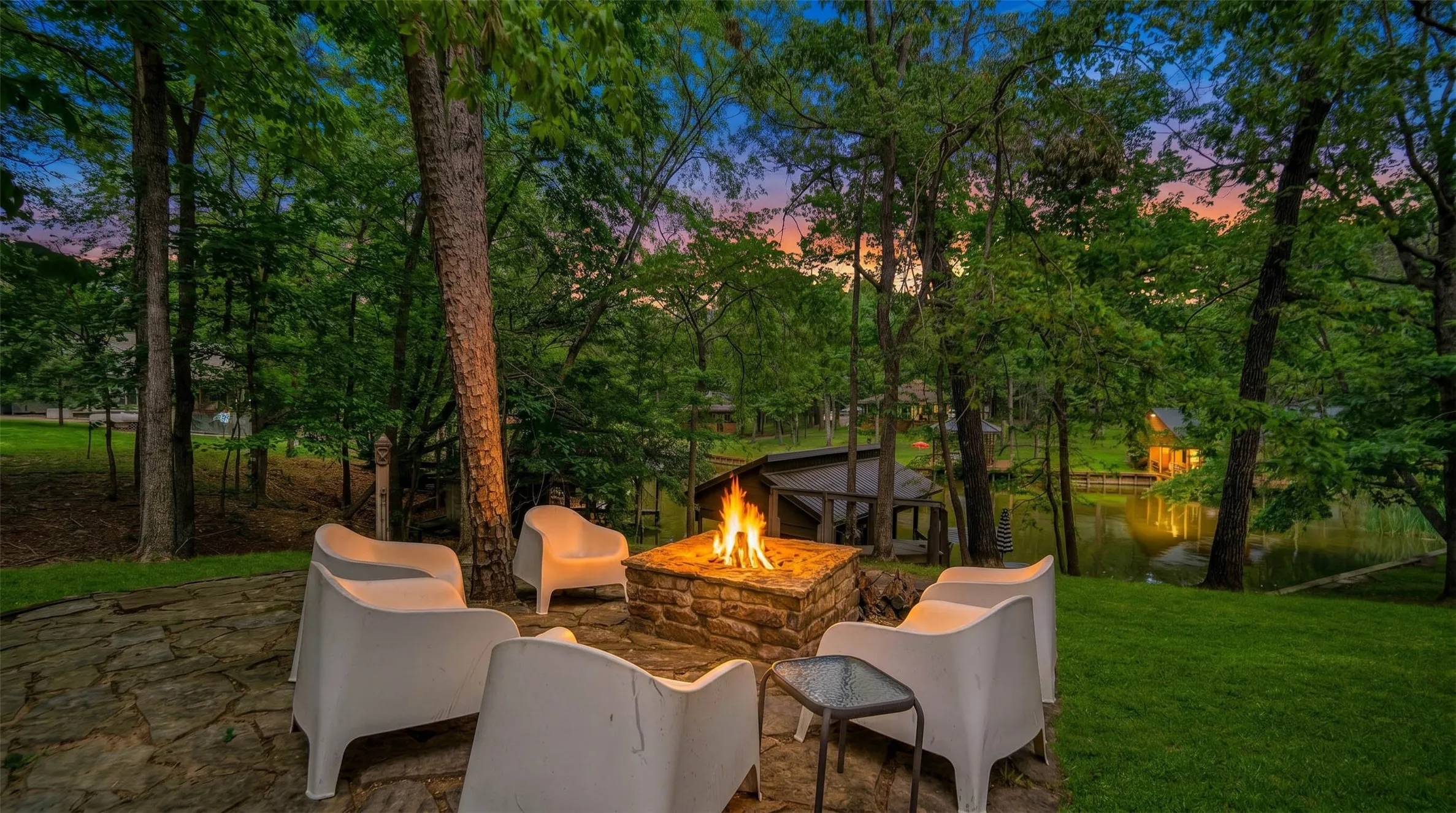 Stone patio with integrated fire pit, expansive lawn, mature tree coverage, and pond frontage