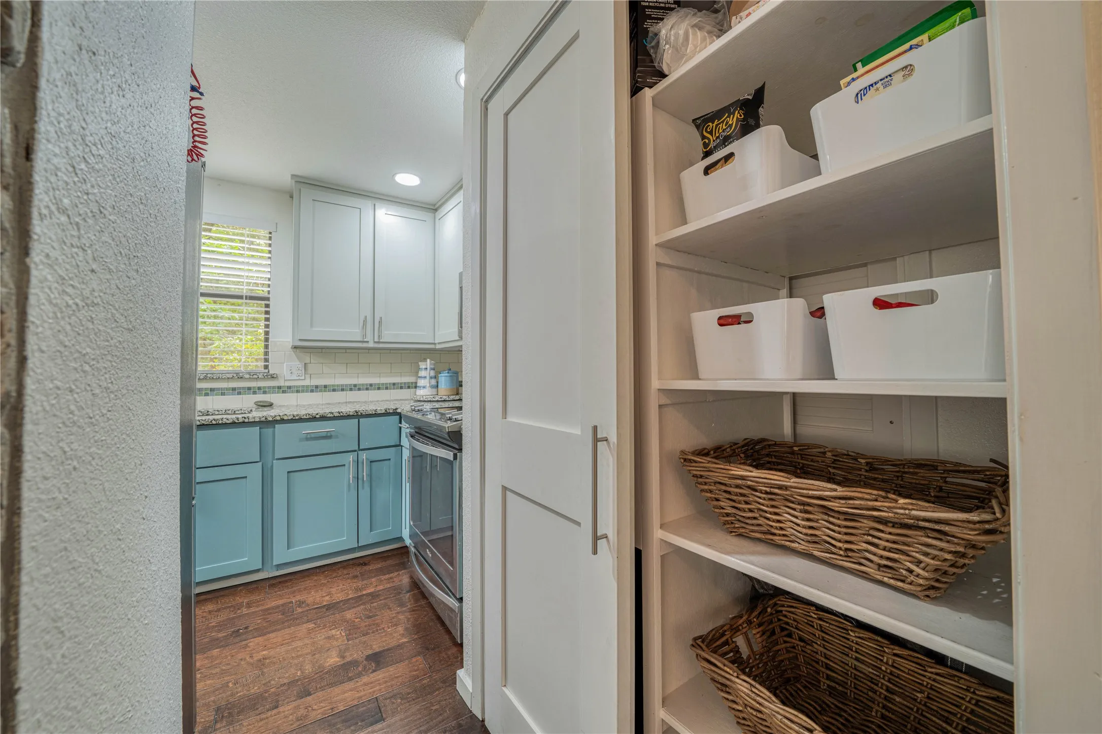 Built-in pantry shelving provides functional storage, complemented by the adjacent kitchen featuring wood-finish flooring, two-tone cabinetry, a tile backsplash, and a window with blinds
