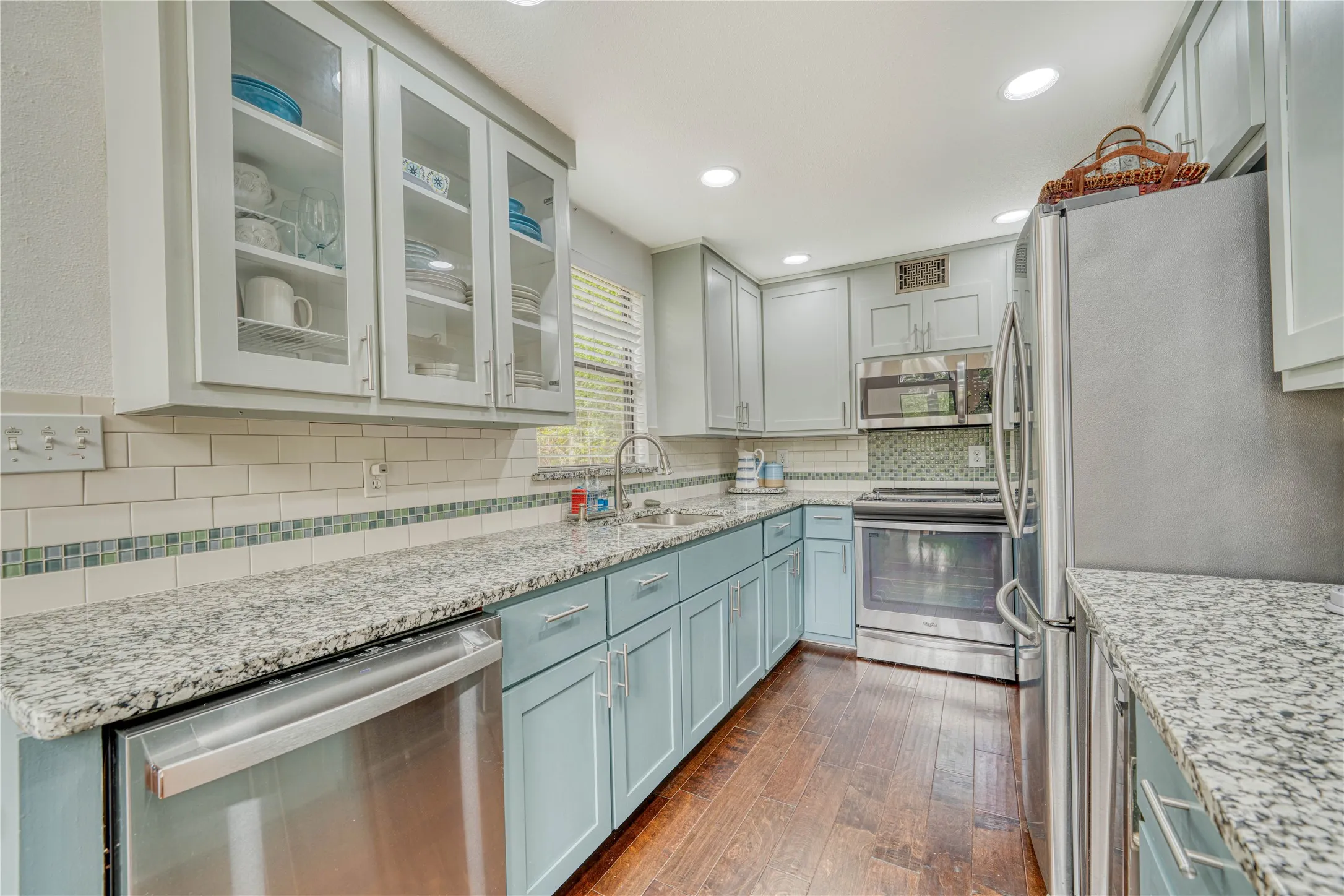 Kitchen featuring blue cabinetry, granite countertops, stainless steel appliances, wood-finish flooring, and subway tile backsplash with decorative accents