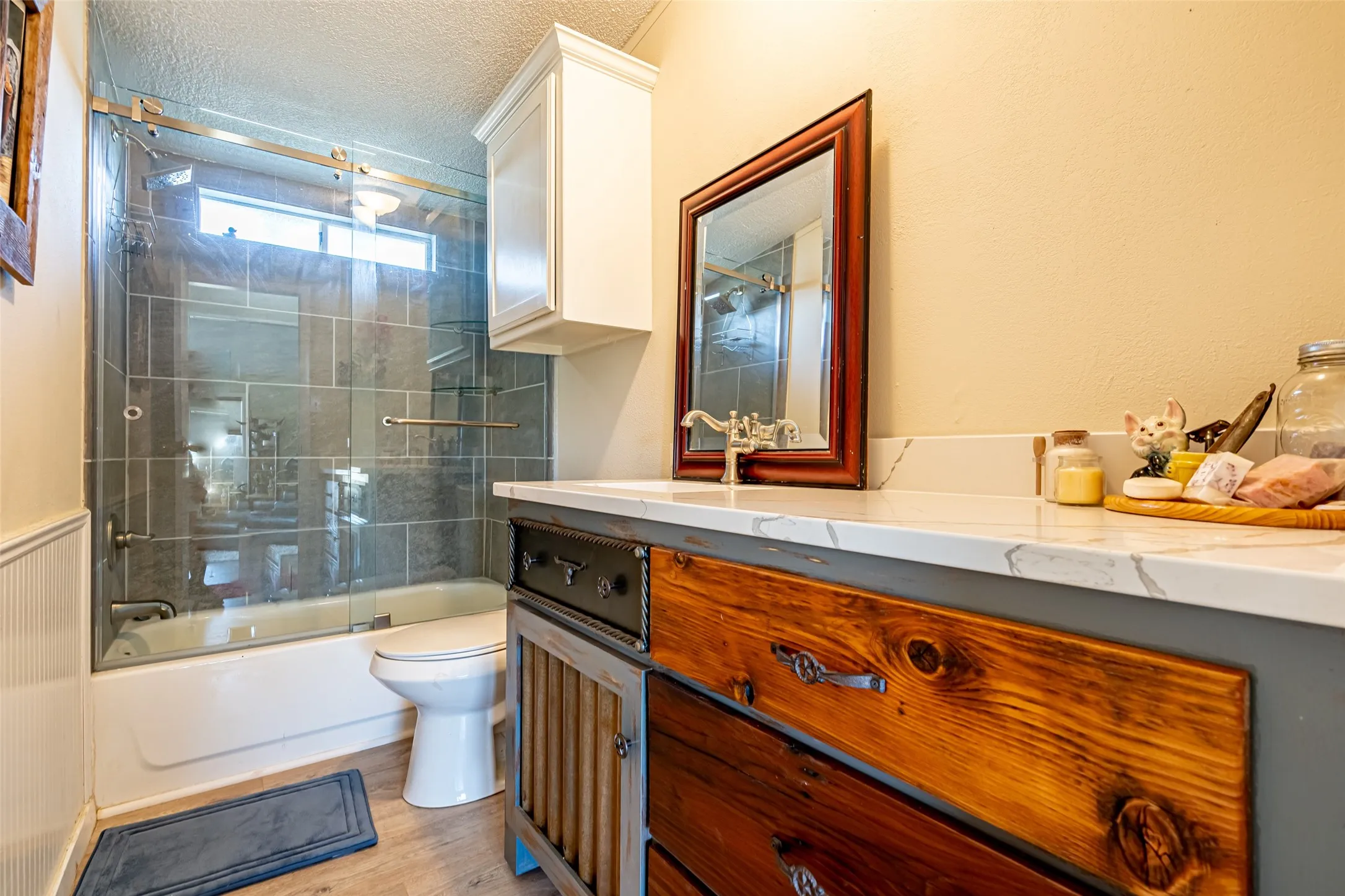 Full bathroom featuring bath / shower combo with glass door, vanity, a textured ceiling, light wood-type flooring, and wainscoting.